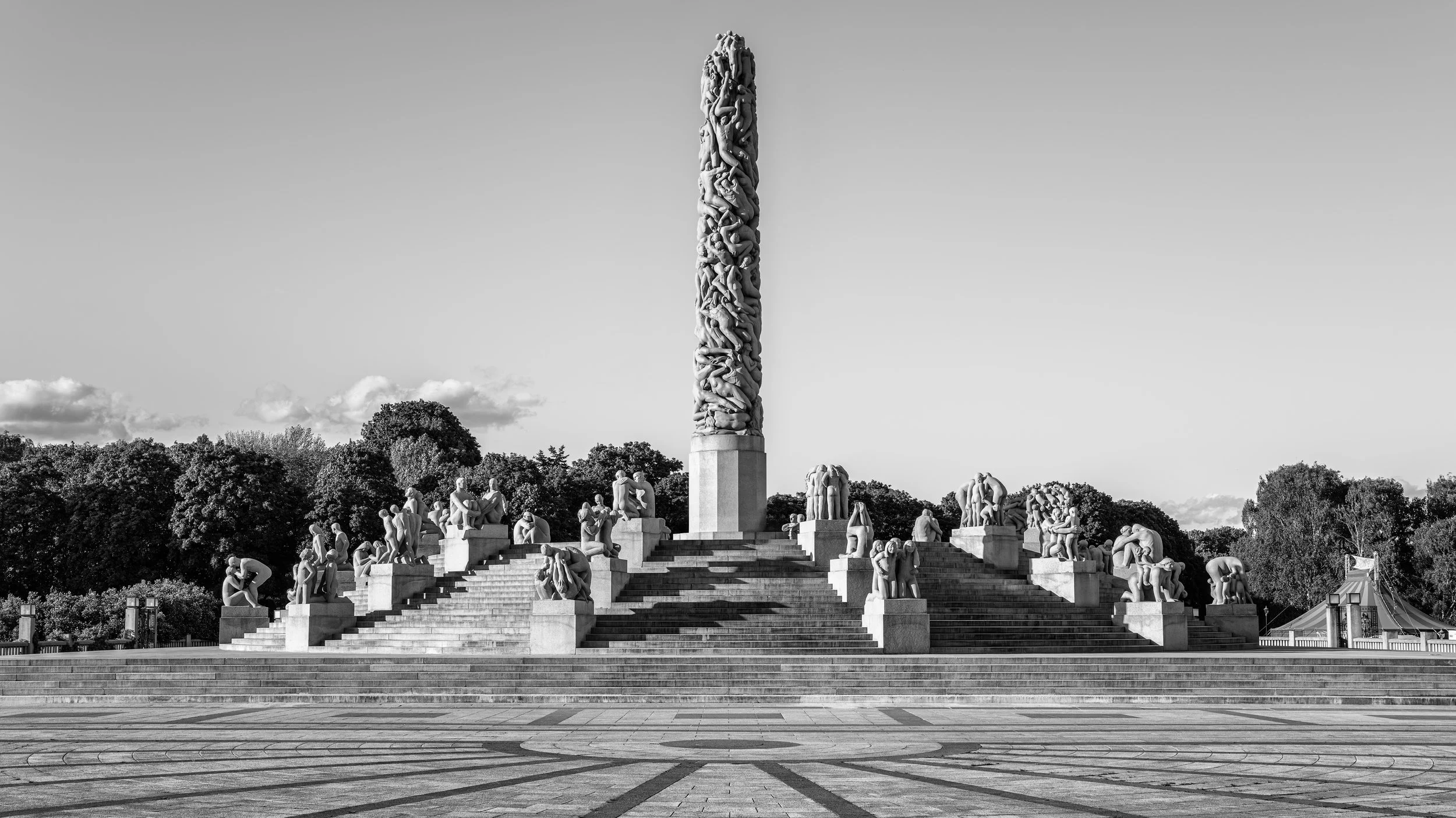 A large monument with numerous human sculptures in a park setting, with stairs leading up to the monument. The sky is mostly clear with a few clouds.