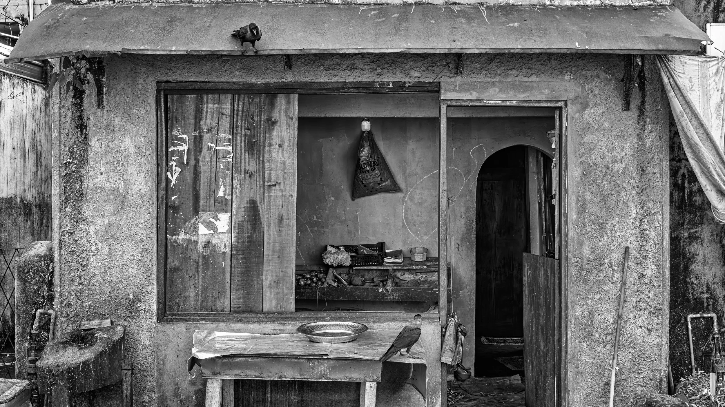 A black and white photo of an old, small building with a weathered appearance. The front has an open doorway and an open window with a hanging bag inside. There is a small shelf with various items including fruits and papers. A wire basket and some o