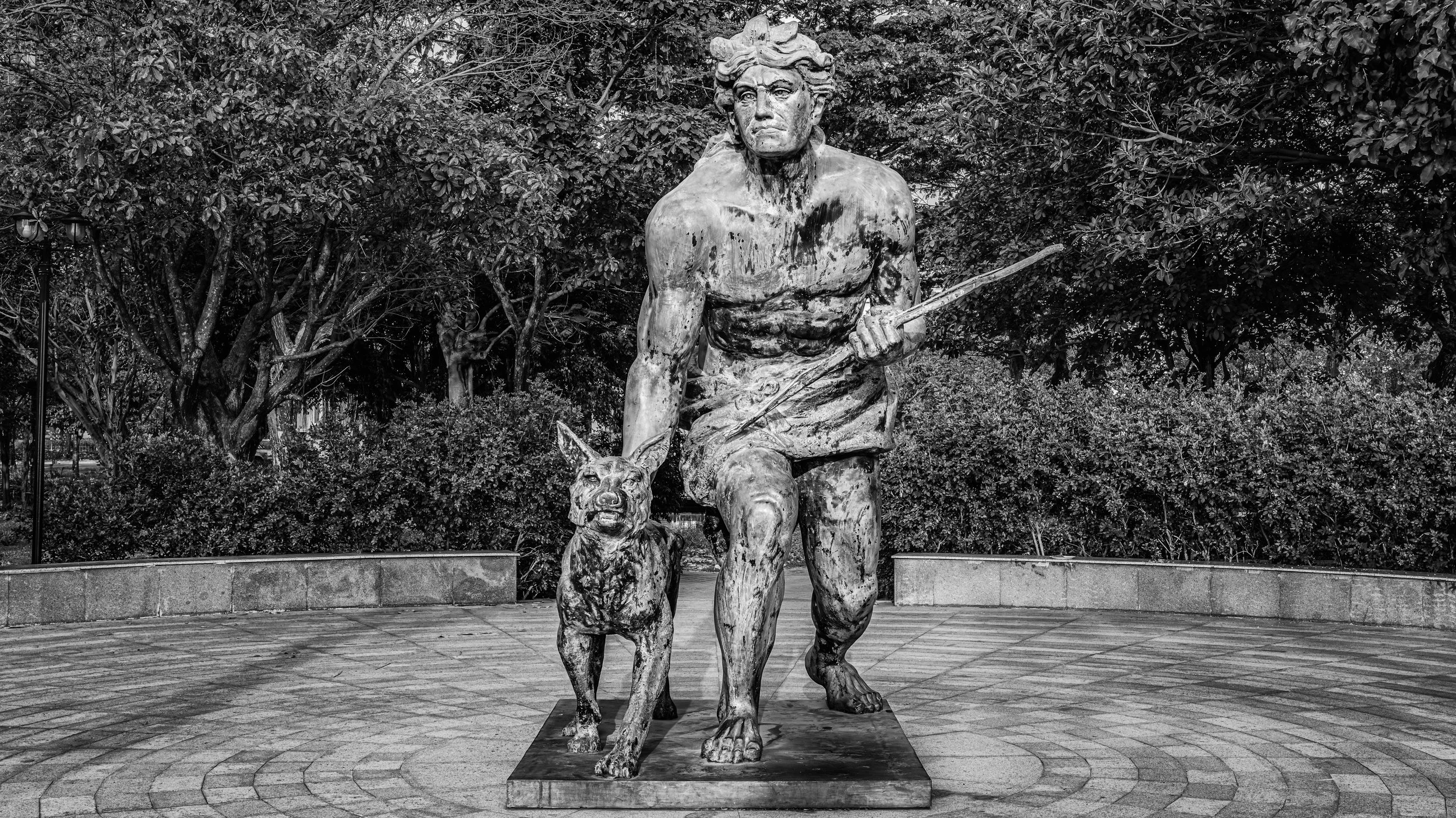 A sculpture of a muscular man with a stern expression, holding a spear in his right hand, with a dog at his feet, set against a backdrop of trees in a park.