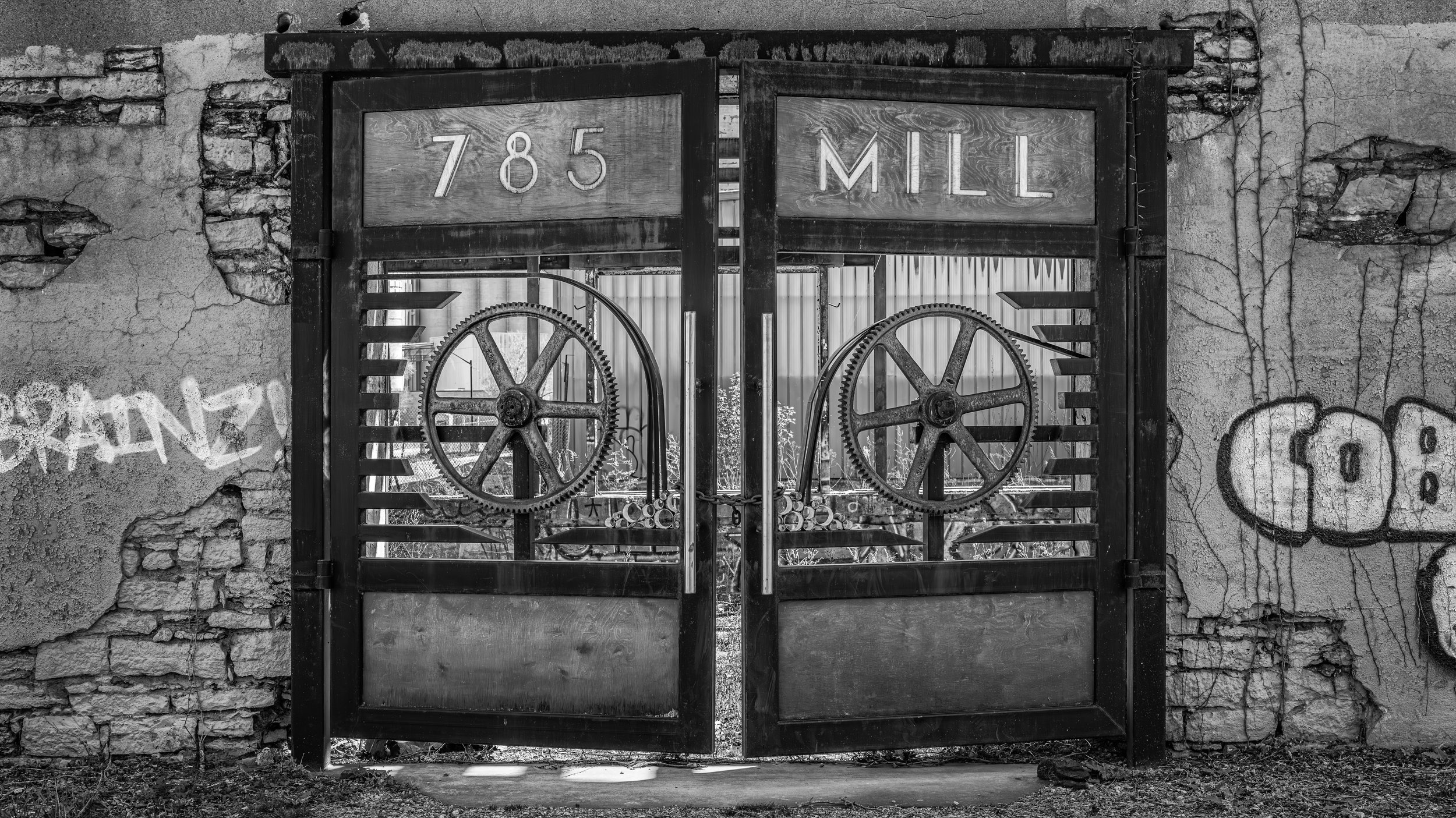 An old, wrought iron gate with gears and cogs as decoration, set in a brick and concrete wall, with a sign that reads '785 MILL' at the top. The scene is in black and white.