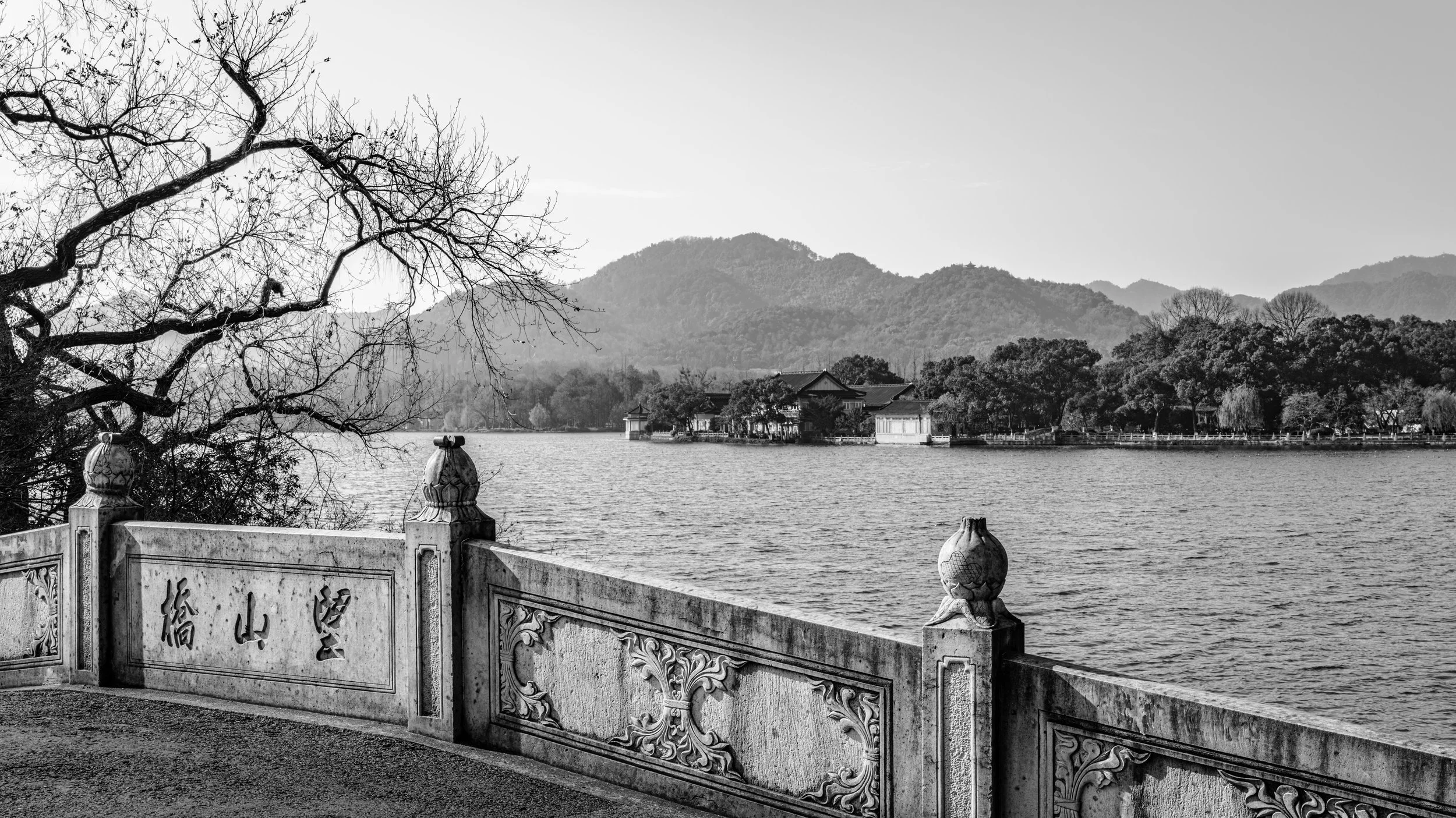 Black and white photo of a lake with a traditional Chinese-style stone railing in the foreground, mountains and trees in the background, and a leafless tree on the left side.