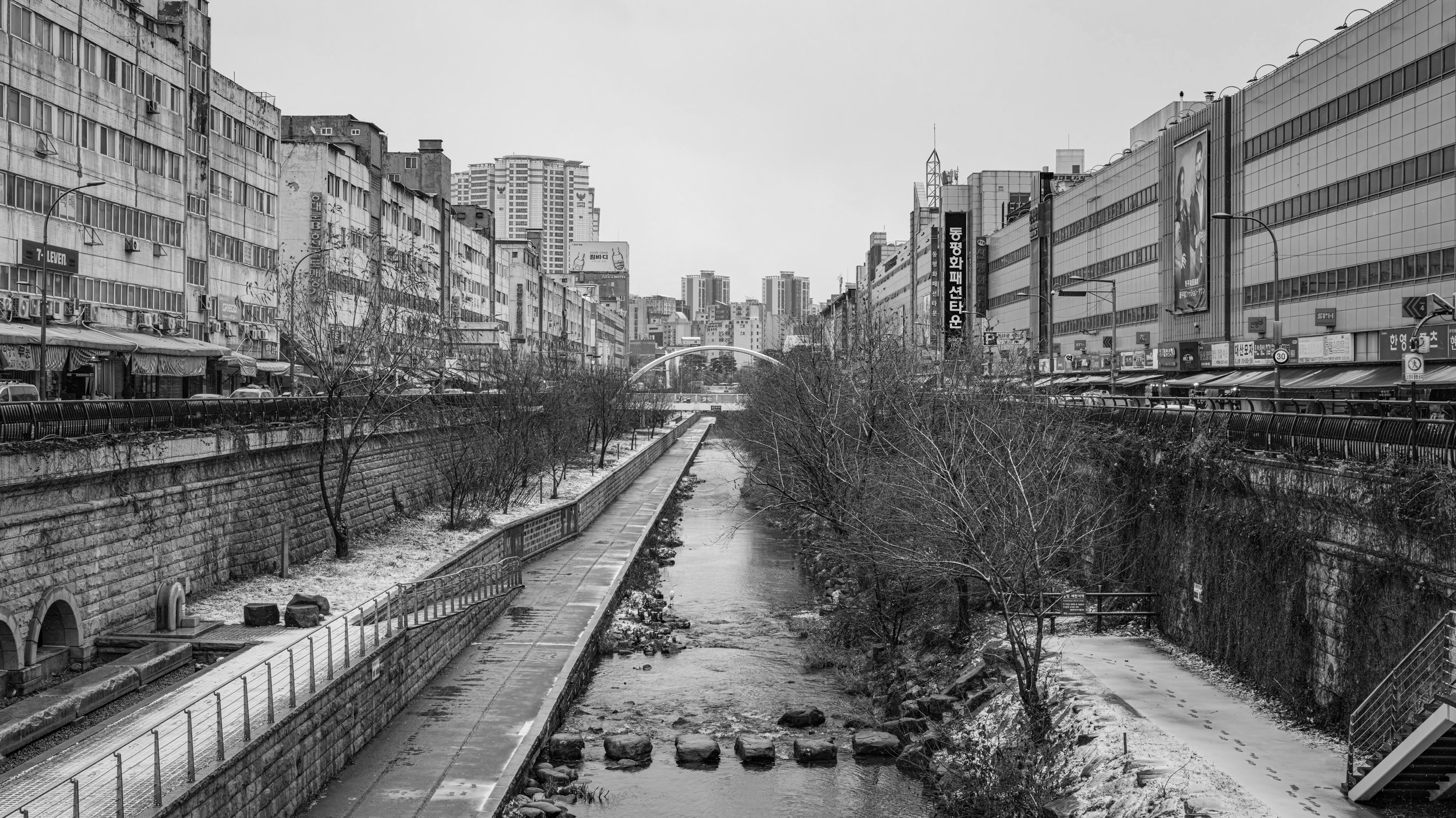 Urban cityscape with tall buildings on both sides of a dry, narrow river or canal, overcast sky, some trees and street signs, black and white photograph.