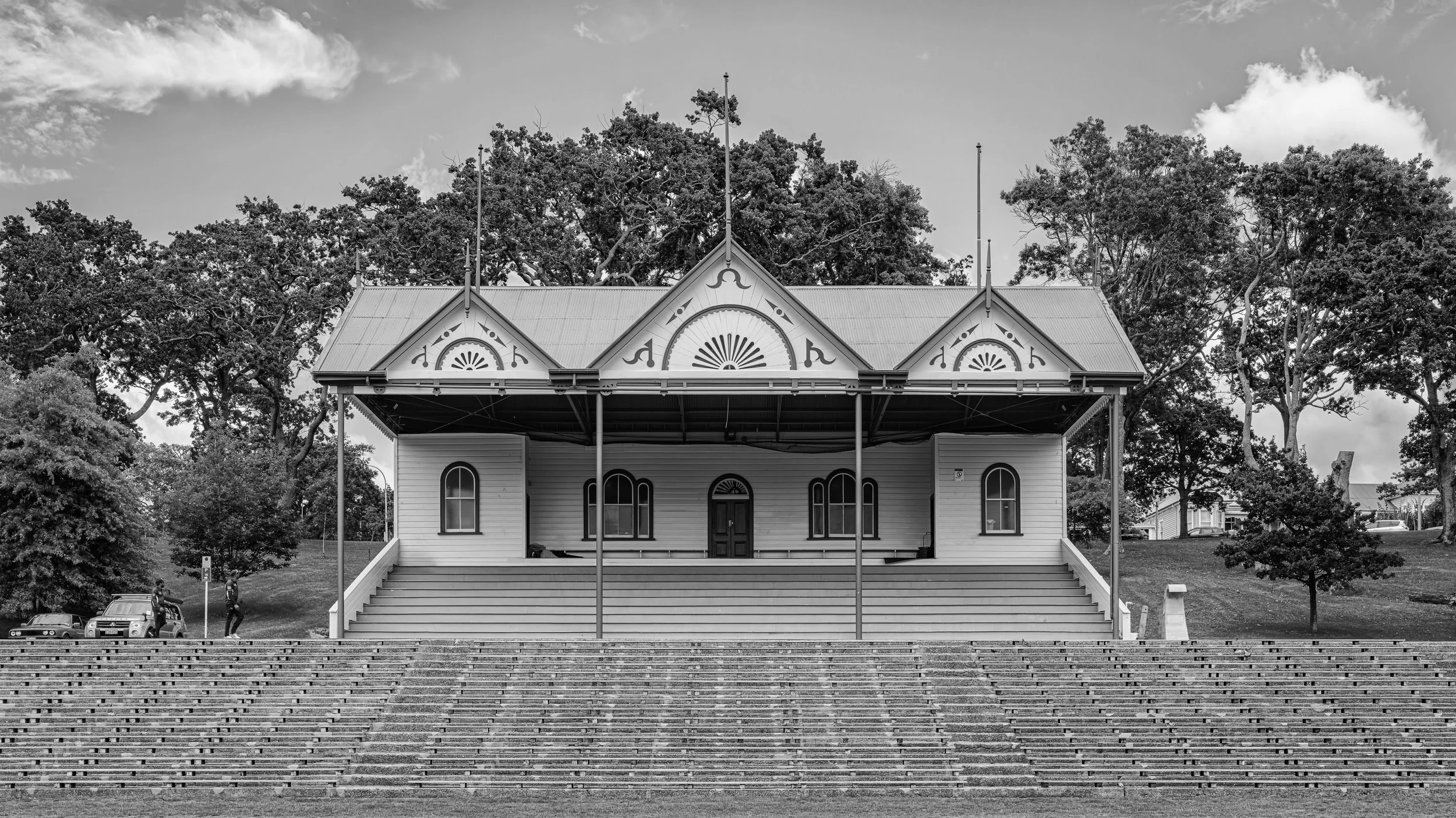 A historic, white wooden building with ornate architectural details and arched windows, situated on a hill with stairs leading up to it. Tall trees surround the building, and a few cars and people are visible in the background. The image is in black 