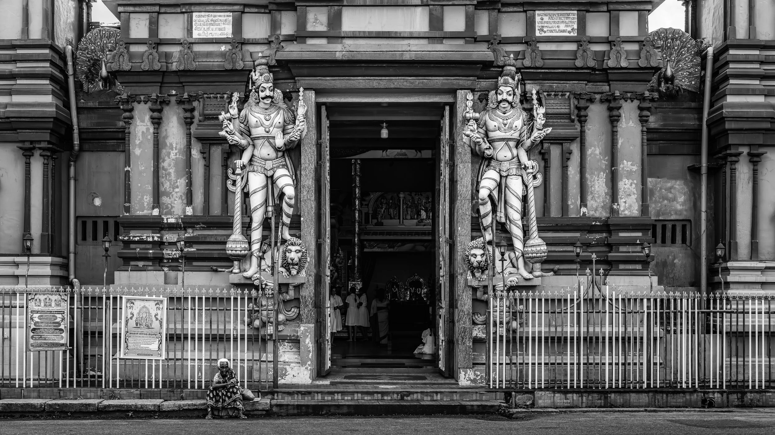 Black and white photo of the entrance to a temple with large carved statues of deities on either side, surrounded by a metal fence. Several people are visible inside the temple and a woman sitting outside.