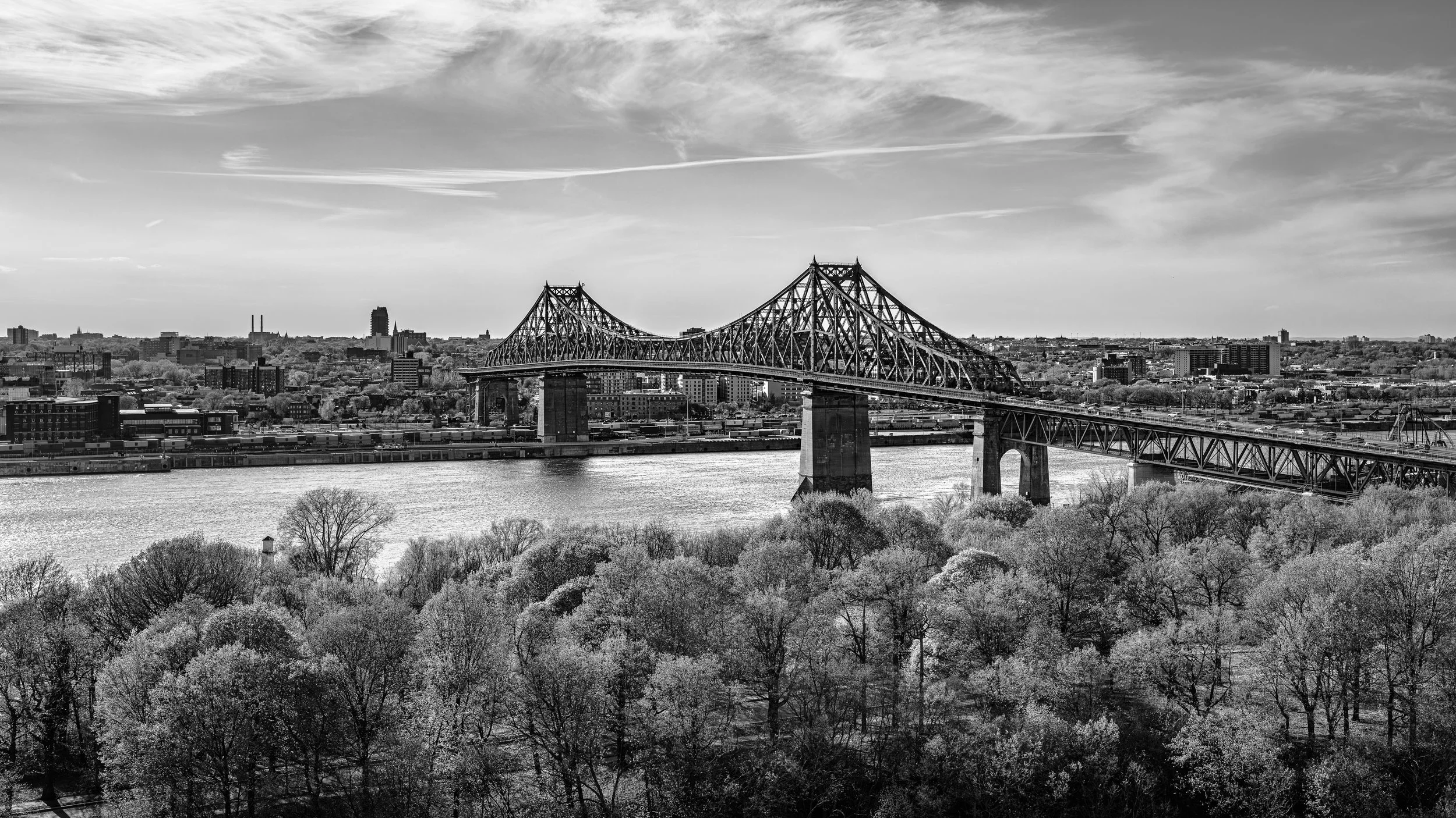 Black and white photo of a city skyline with a large bridge crossing a river, trees in the foreground, and a partly cloudy sky.