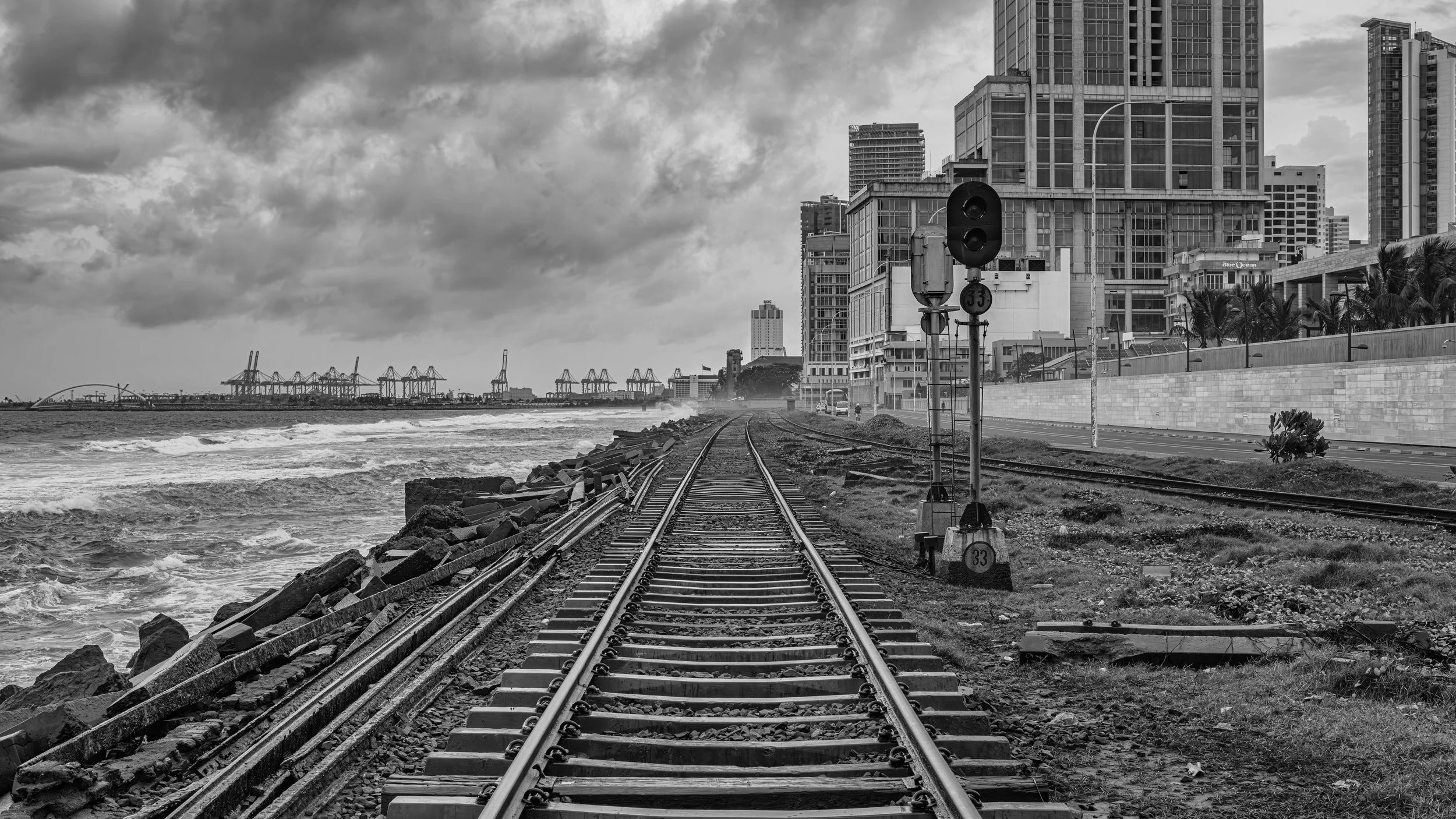 Black and white photo of railroad tracks running along a city waterfront with high-rise buildings on the right and shipping cranes in the distance on the left under a cloudy sky.