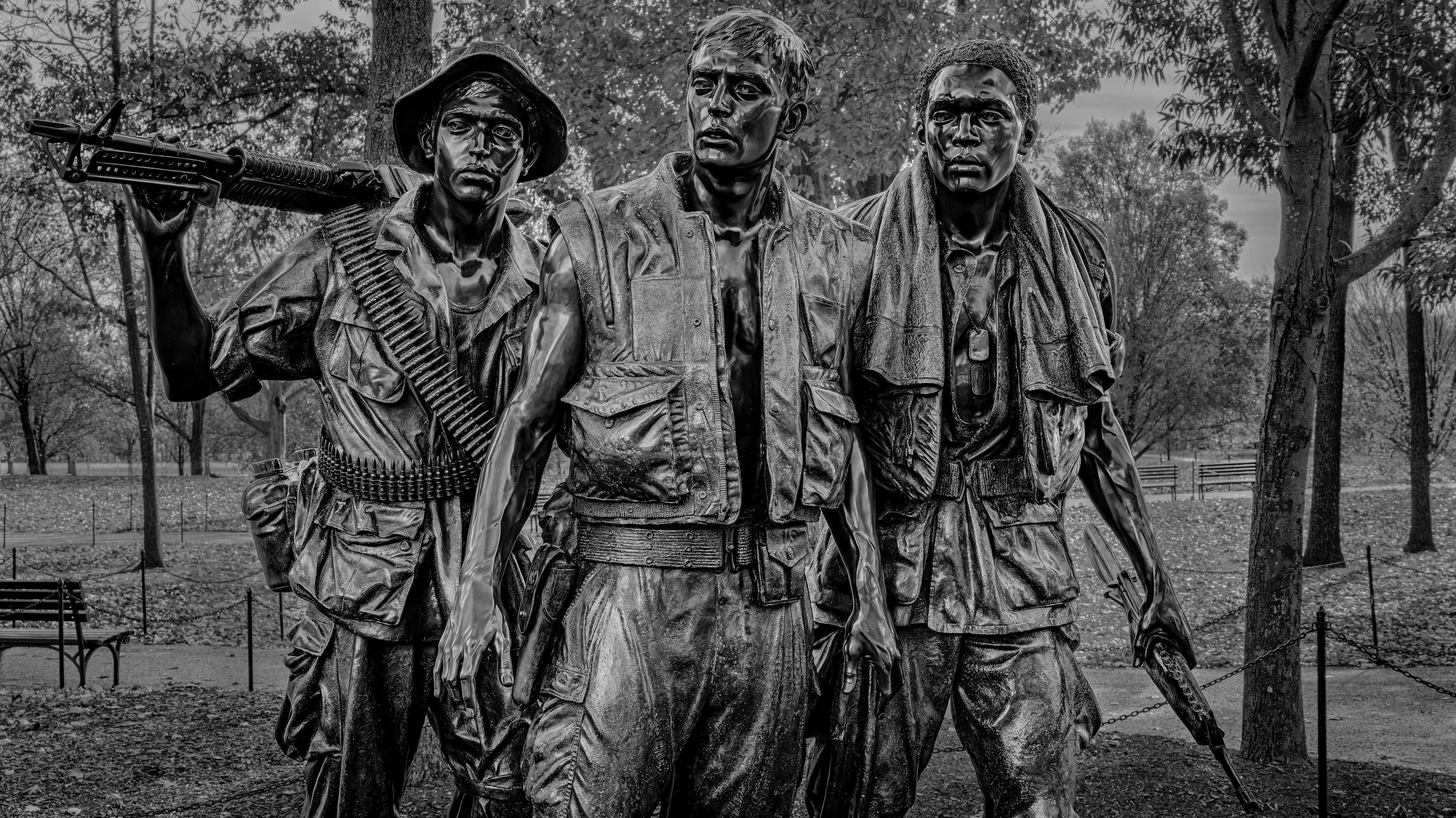 A black and white statue of three soldiers in military gear, standing outdoors with trees in the background.