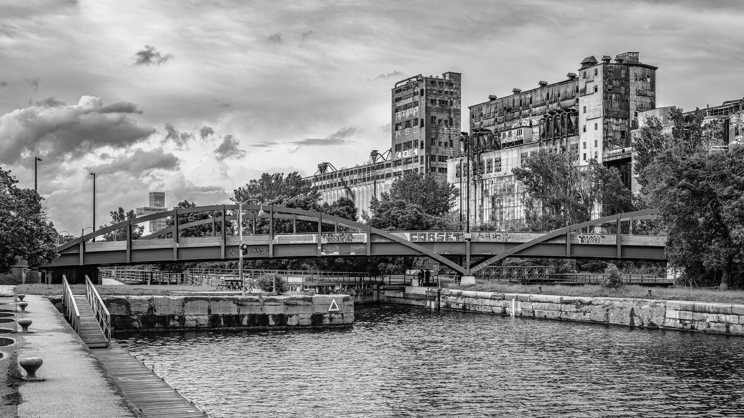 Black and white photo of an abandoned industrial building near a waterway with a bridge overhead and trees in the foreground.