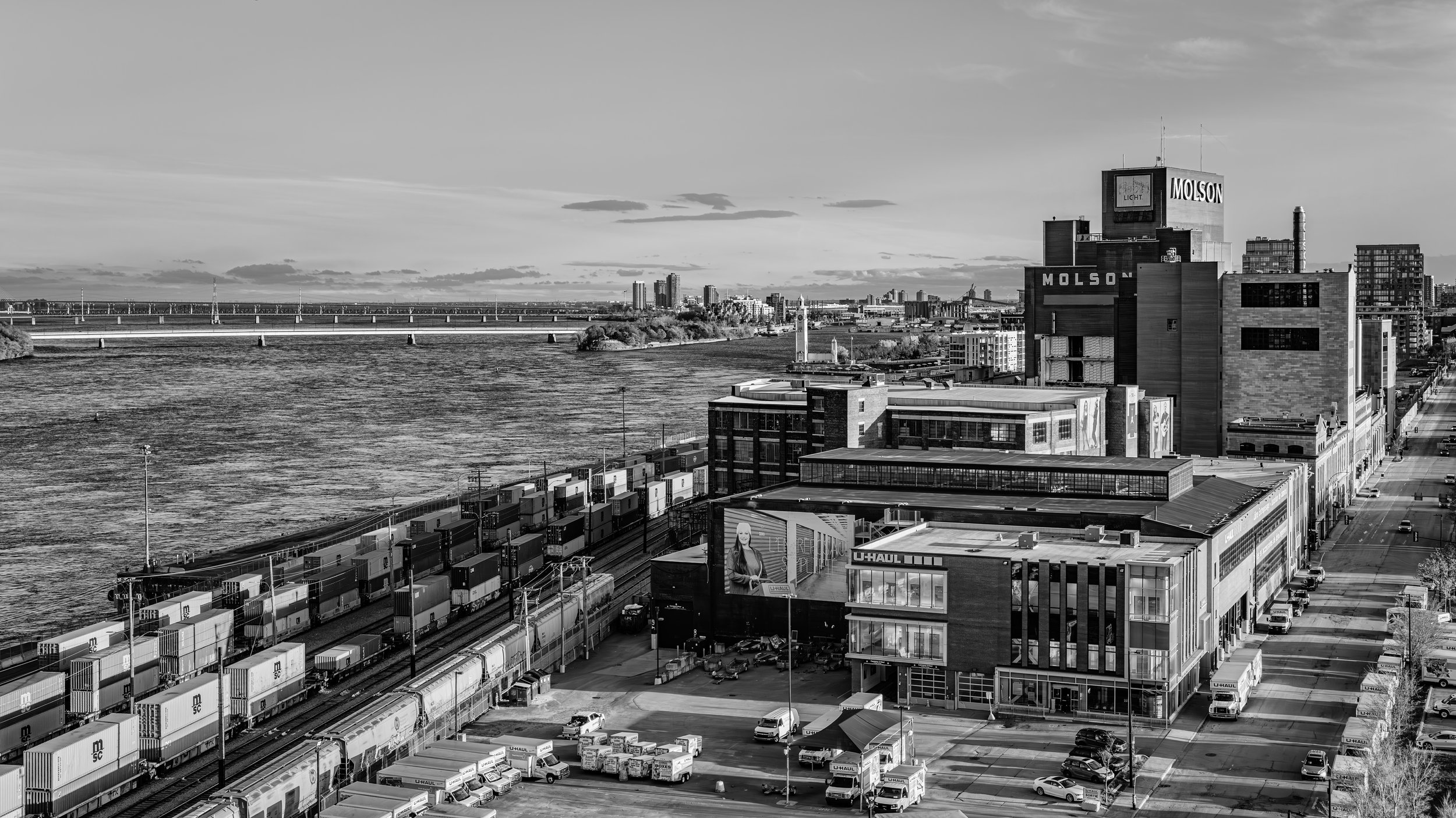 Black and white photo of a cityscape with a river, a bridge, and tall buildings. In the foreground, there are U-Haul trucks and storage containers. On the right, a tall building displays the words 'MOLSON' and 'LIGHT.'
