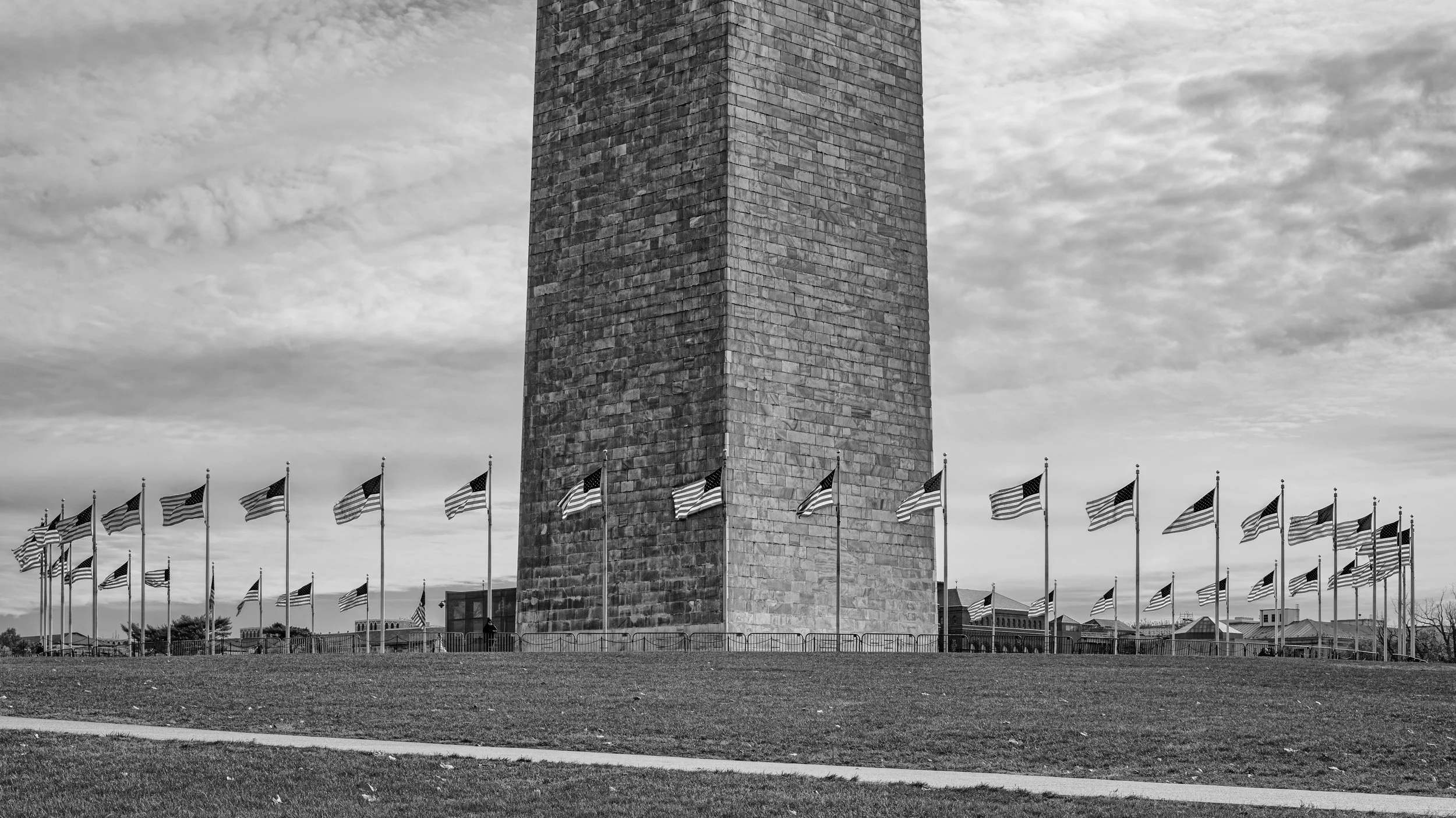 A large stone monument known as the Washington Monument, surrounded by United States flags flying on flagpoles, under a cloudy sky.