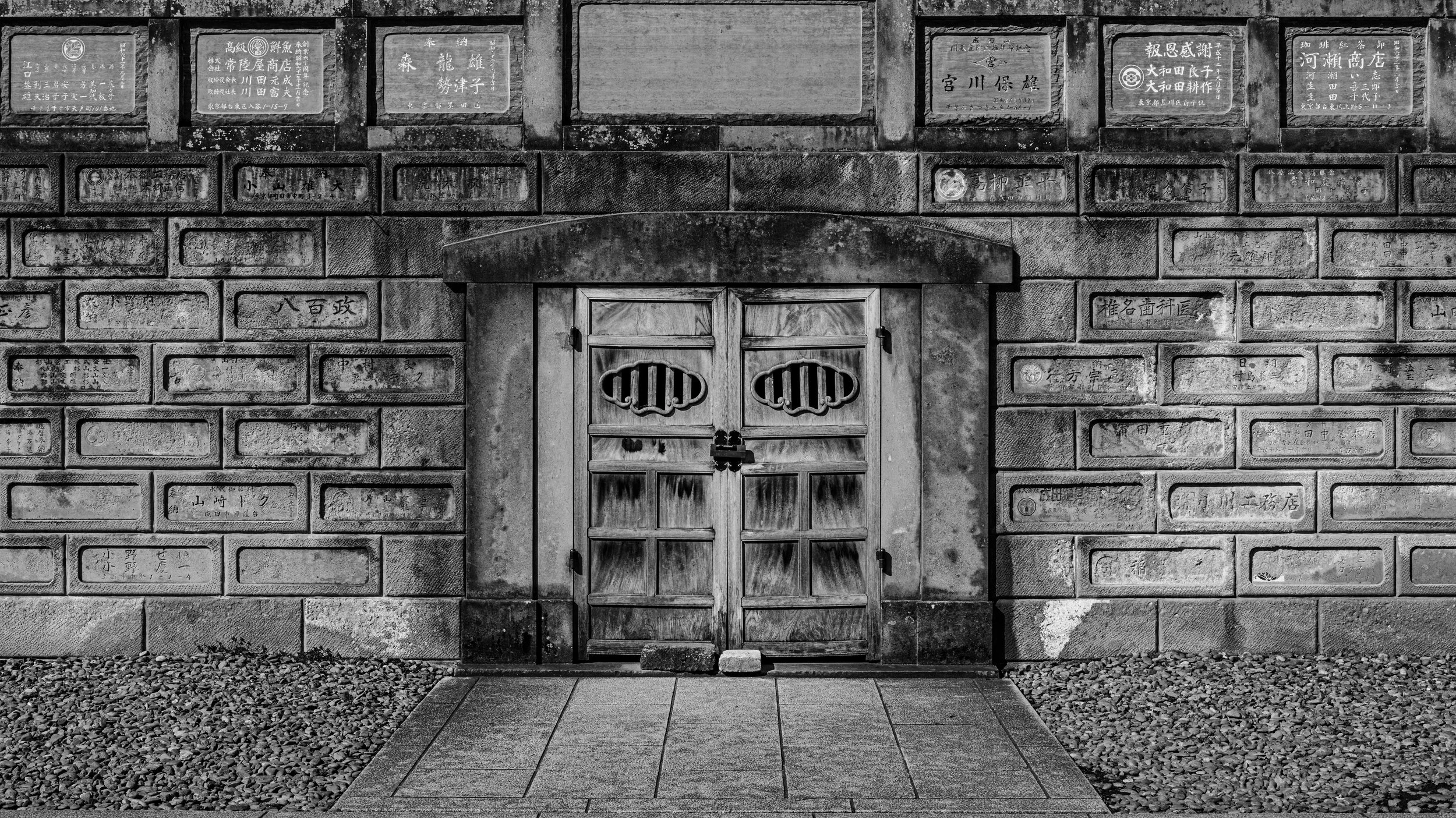 A black and white photo of a traditional Japanese building with a stone wall and a wooden door with ornate carvings. The wall is decorated with numerous engraved plaques with Japanese writing.