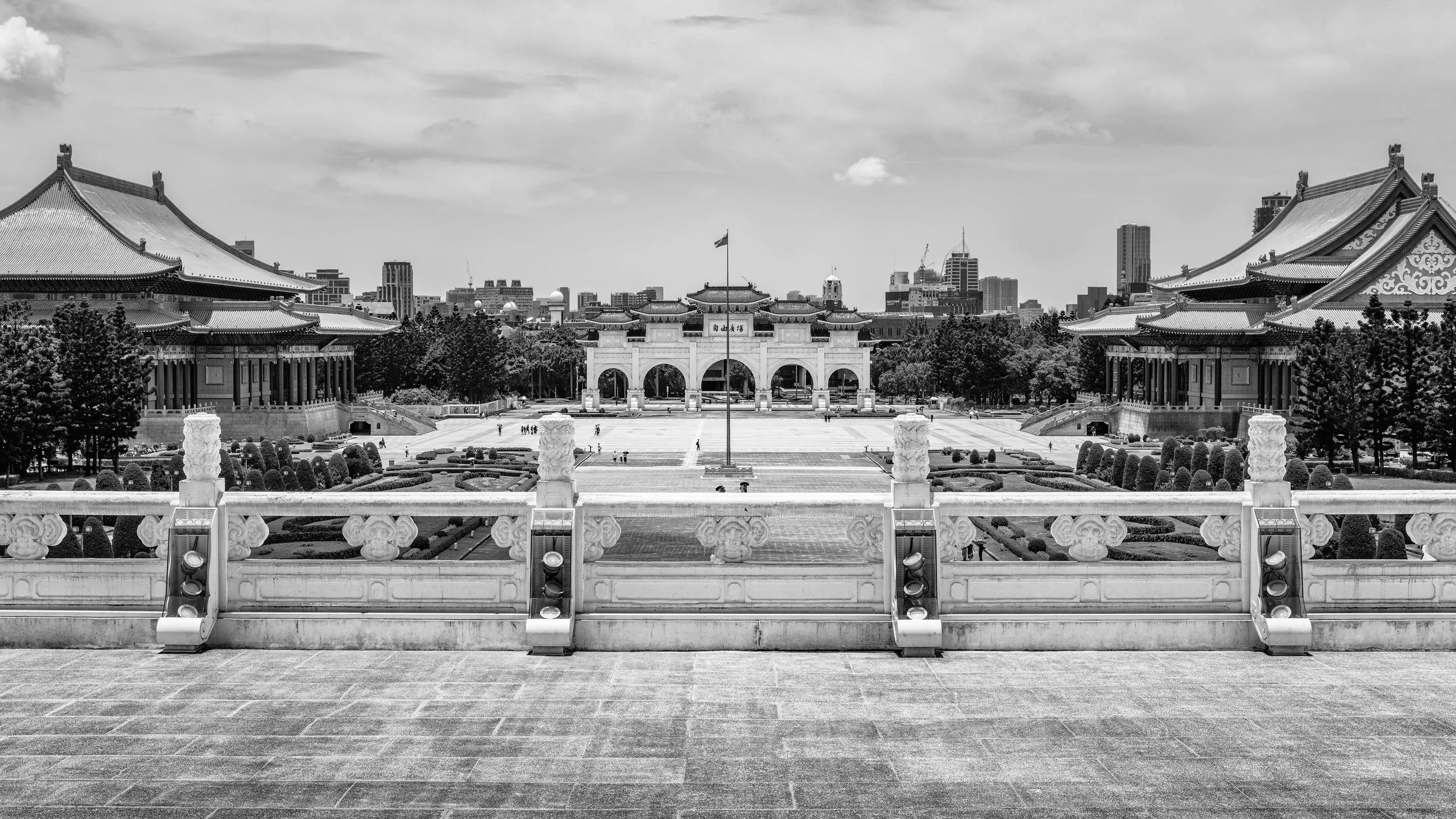 Black and white photo of a large traditional Chinese-style temple with an archway, flanked by two smaller pagoda-style buildings, overlooking a landscaped garden and a city skyline with high-rise buildings in the background.
