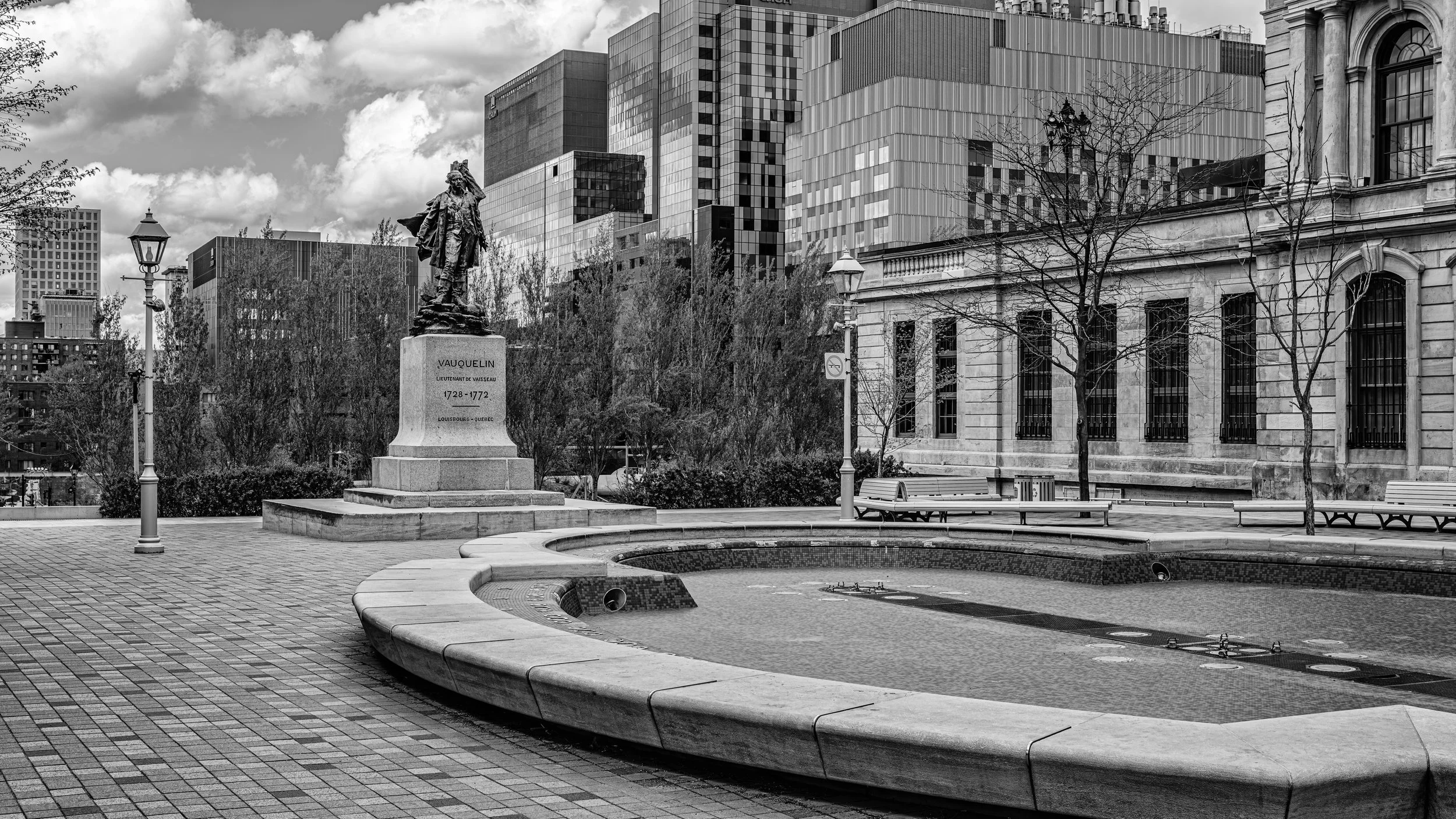 A black and white photo of a city park with a statue of a historical figure, VAUQUELIN, on a pedestal. Surrounding the statue are leafless trees, benches, and street lamps. Modern high-rise buildings are in the background under a partly cloudy sky.