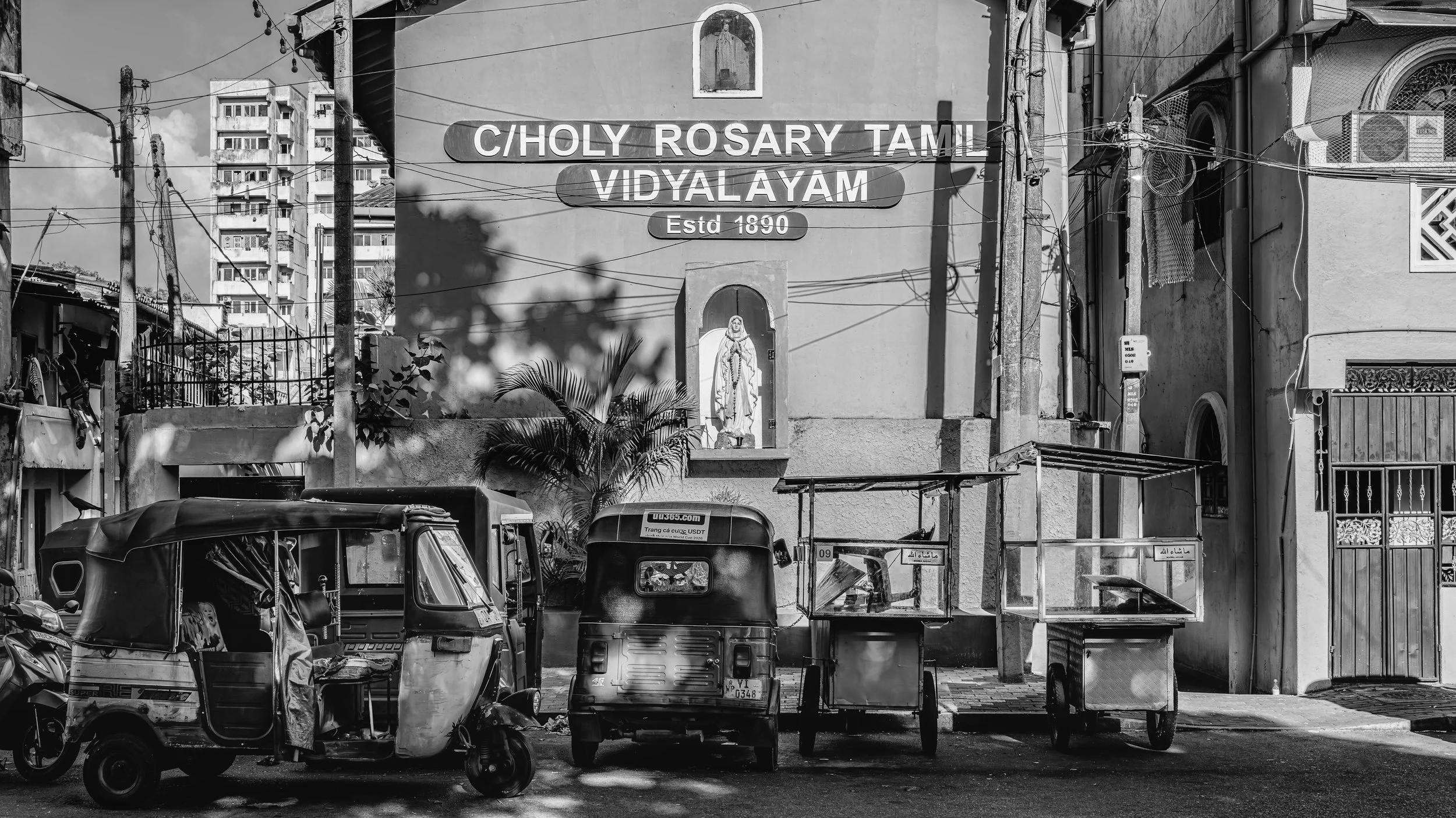 Black and white photo of a street scene with vehicles, a sign reading "C/Choly Rosary Tamil Vidyalayam Estd 1890," and a religious statue under an arch on a building.