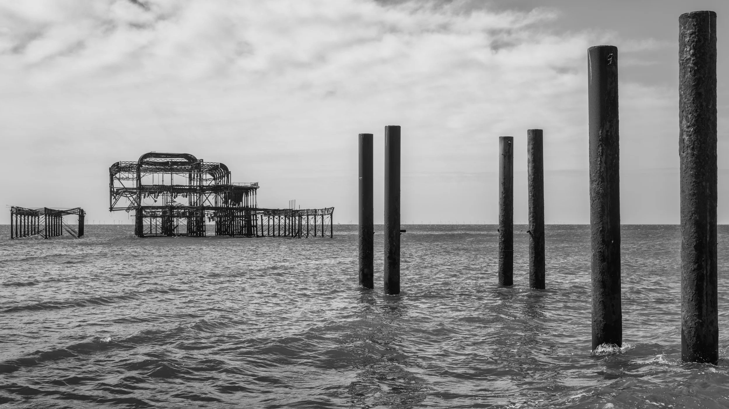 Abandoned and rusted pier structures in the ocean with four large vertical poles in the foreground and partly cloudy sky.