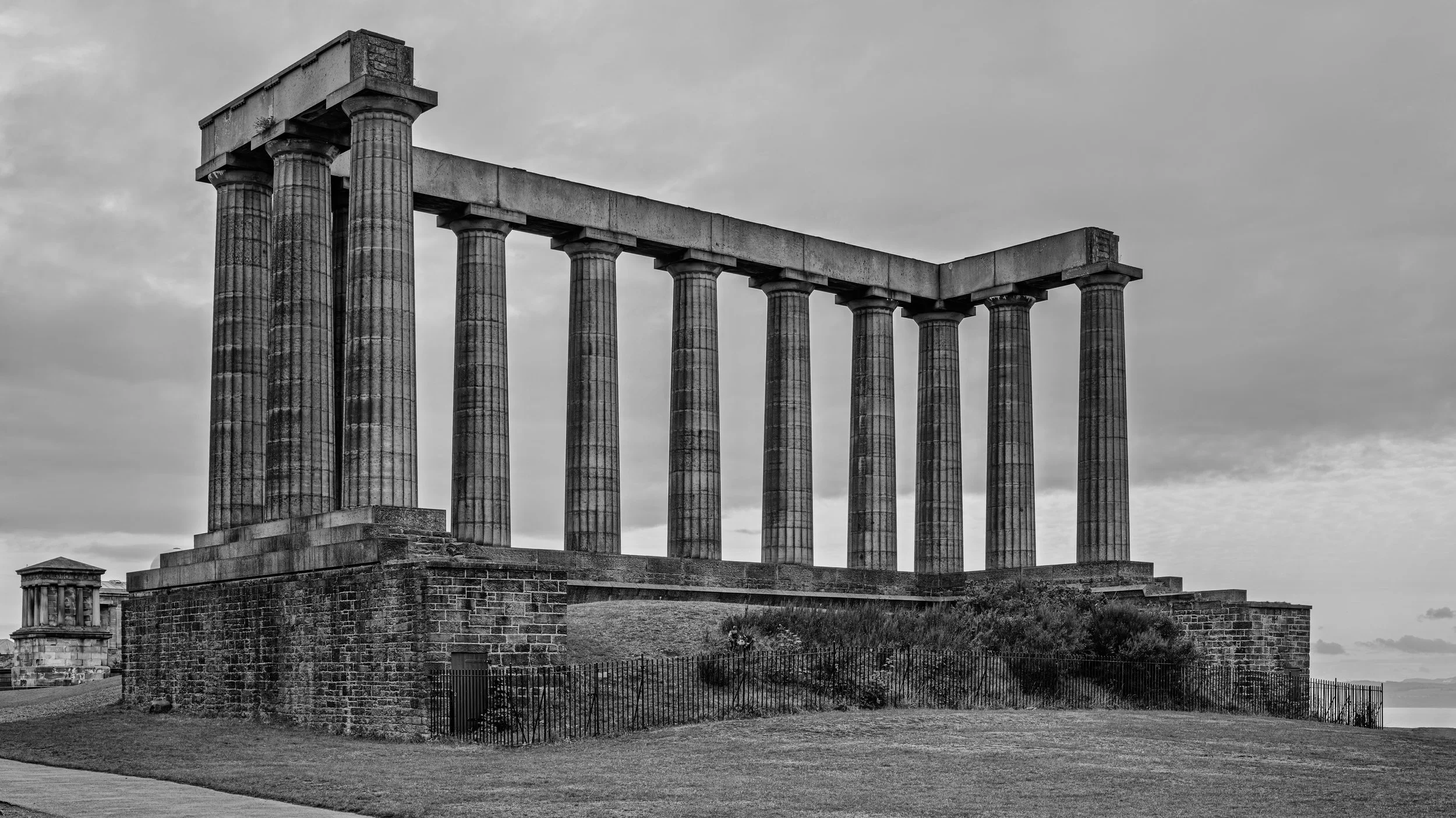Black and white photo of ancient Greek-style ruins with six large columns on a stone platform, a smaller structure with columns in the background, and a cloudy sky.