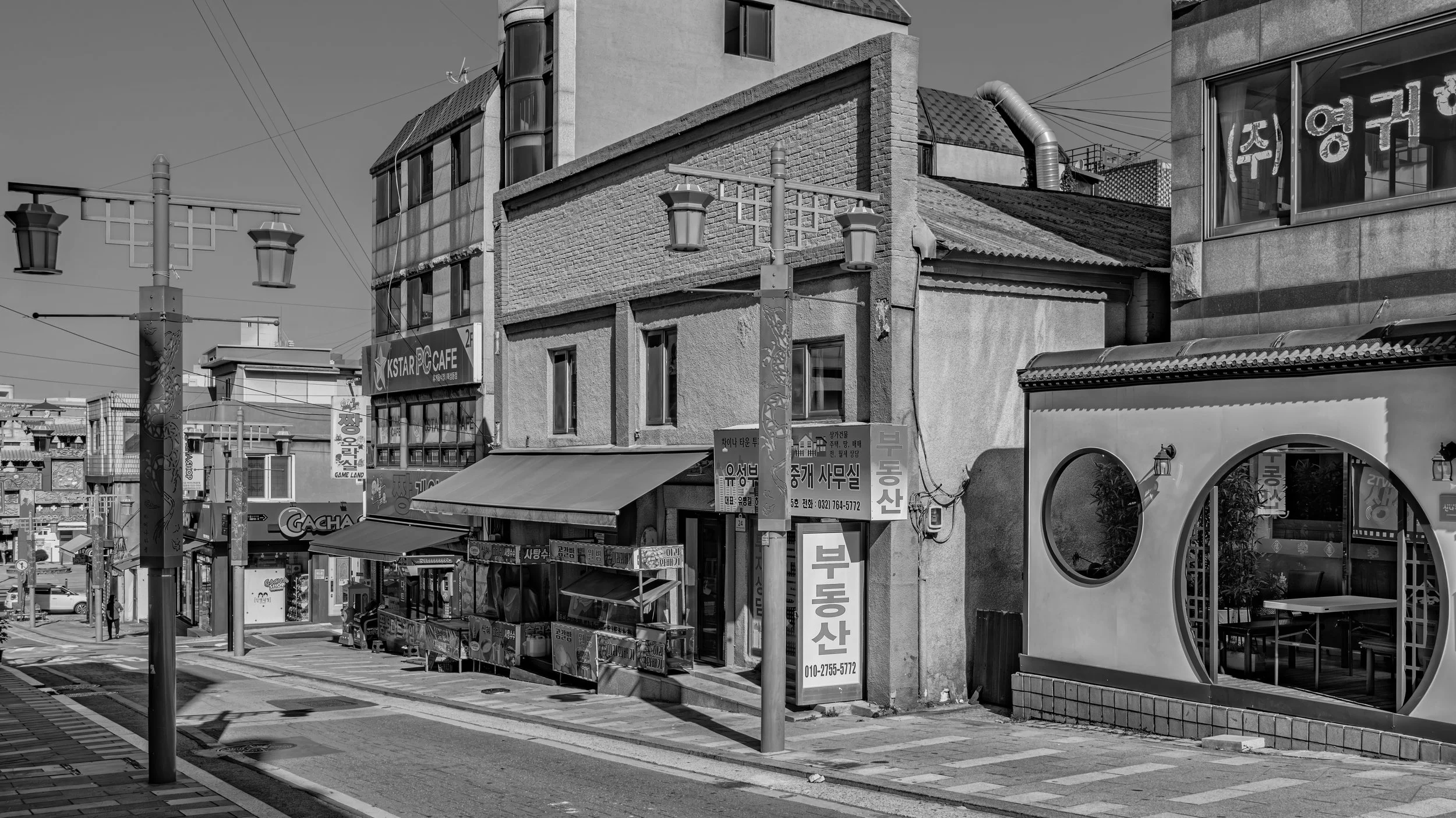 A city street scene with buildings, street lamps, and signage in Korean, viewed in black and white.