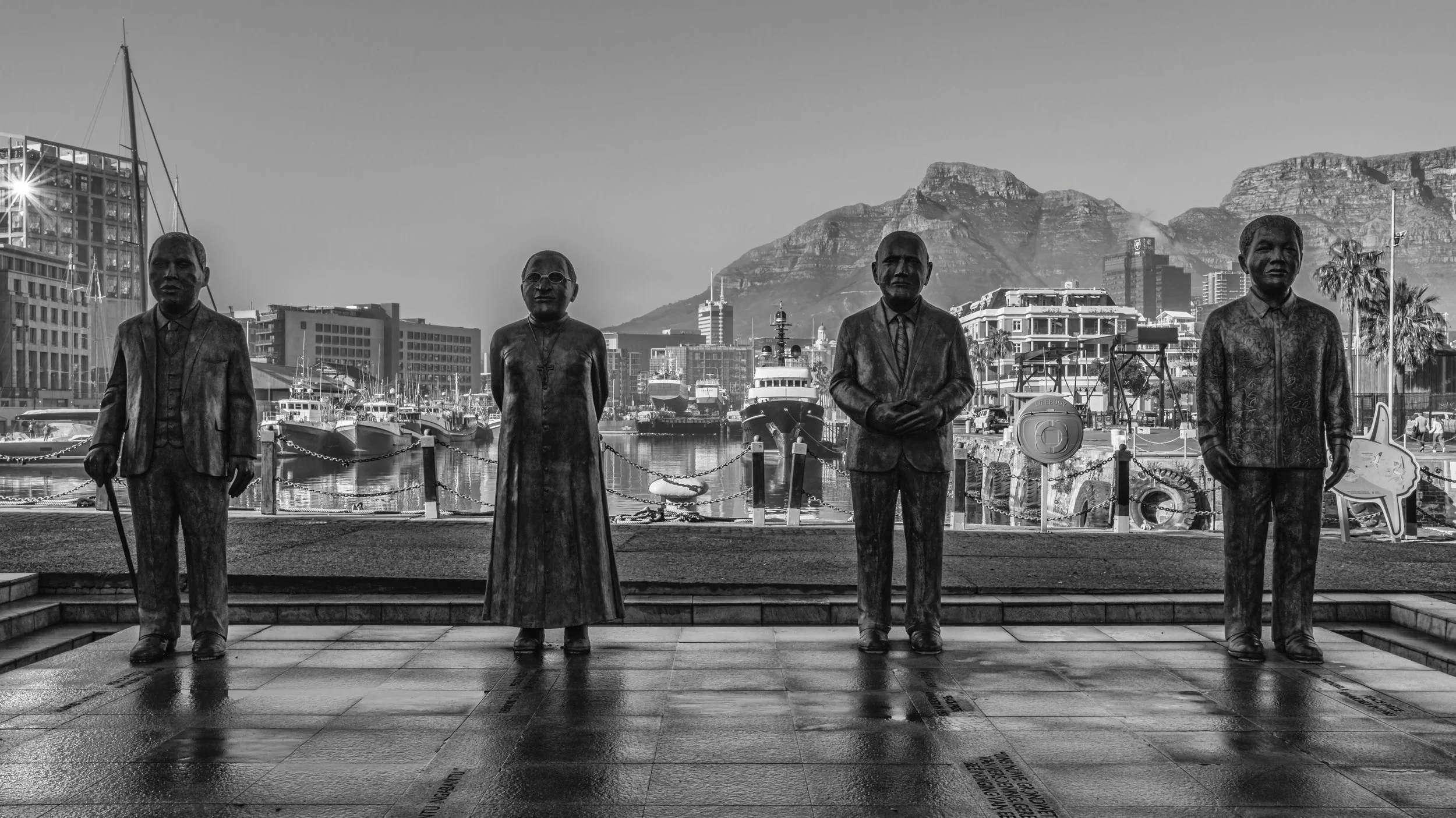 Black and white photo of four bronze statues of historical figures standing by a harbor with boats, city buildings, and mountains in the background. Cape Town, South Africa.
