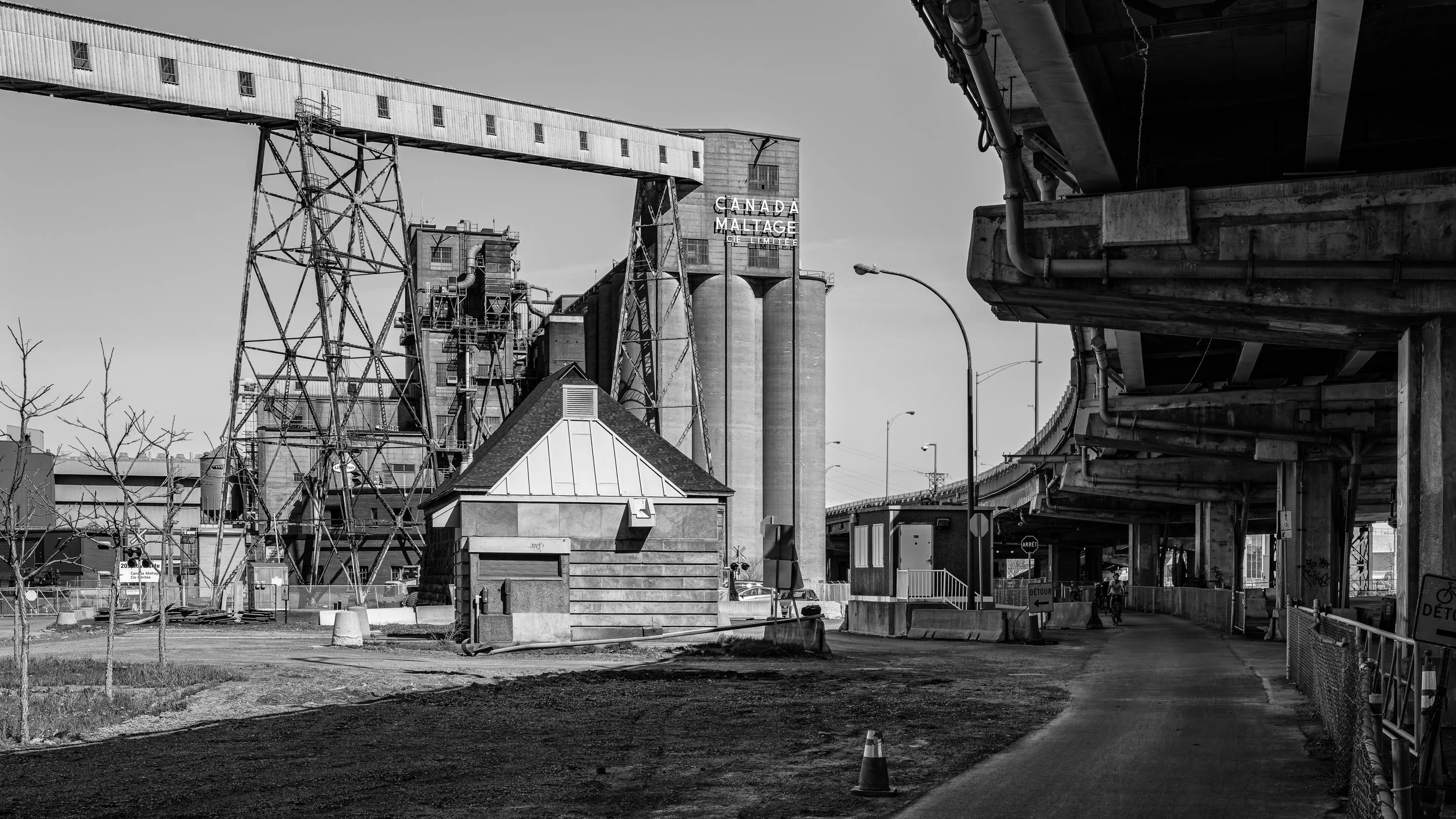 Black and white photo of industrial buildings, silos, a covered bridge, and a street with construction cones and fences.