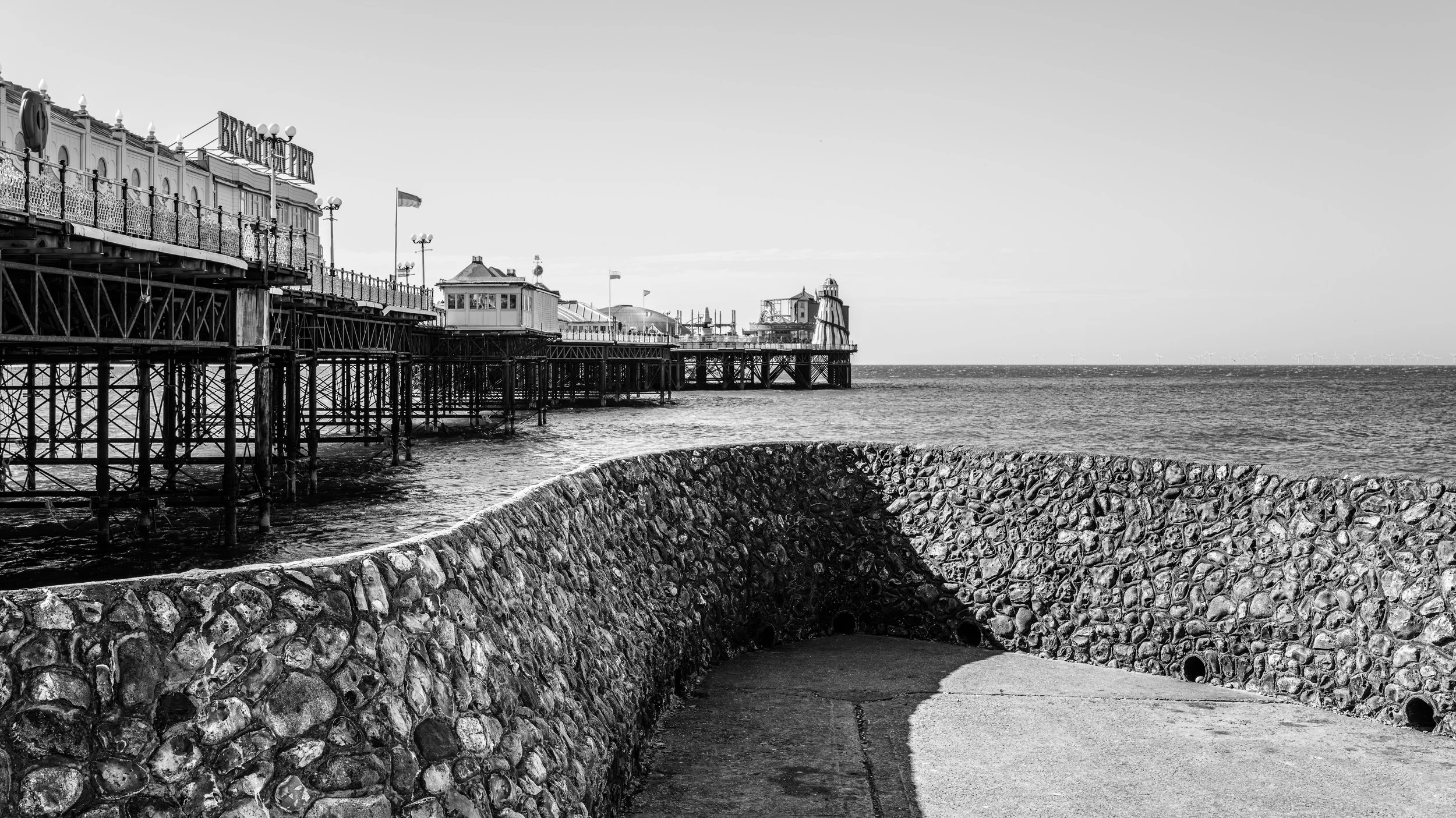 A black and white photograph of a seaside pier extending over water, with a stone embankment in the foreground. The pier has decorative railings, street lamps, and a fixture marked 'Brighton Pier.' There are flags on the pier, and wind turbines are v