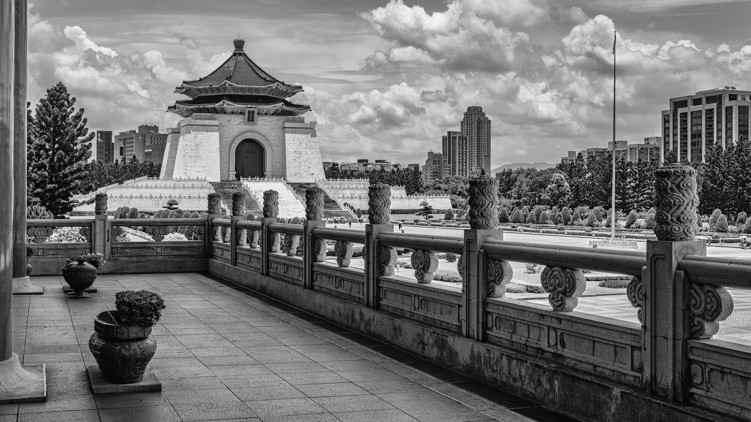 Black and white photo of a traditional East Asian pagoda-style building with ornate roof, set against a modern city skyline with tall buildings and cloudy sky.