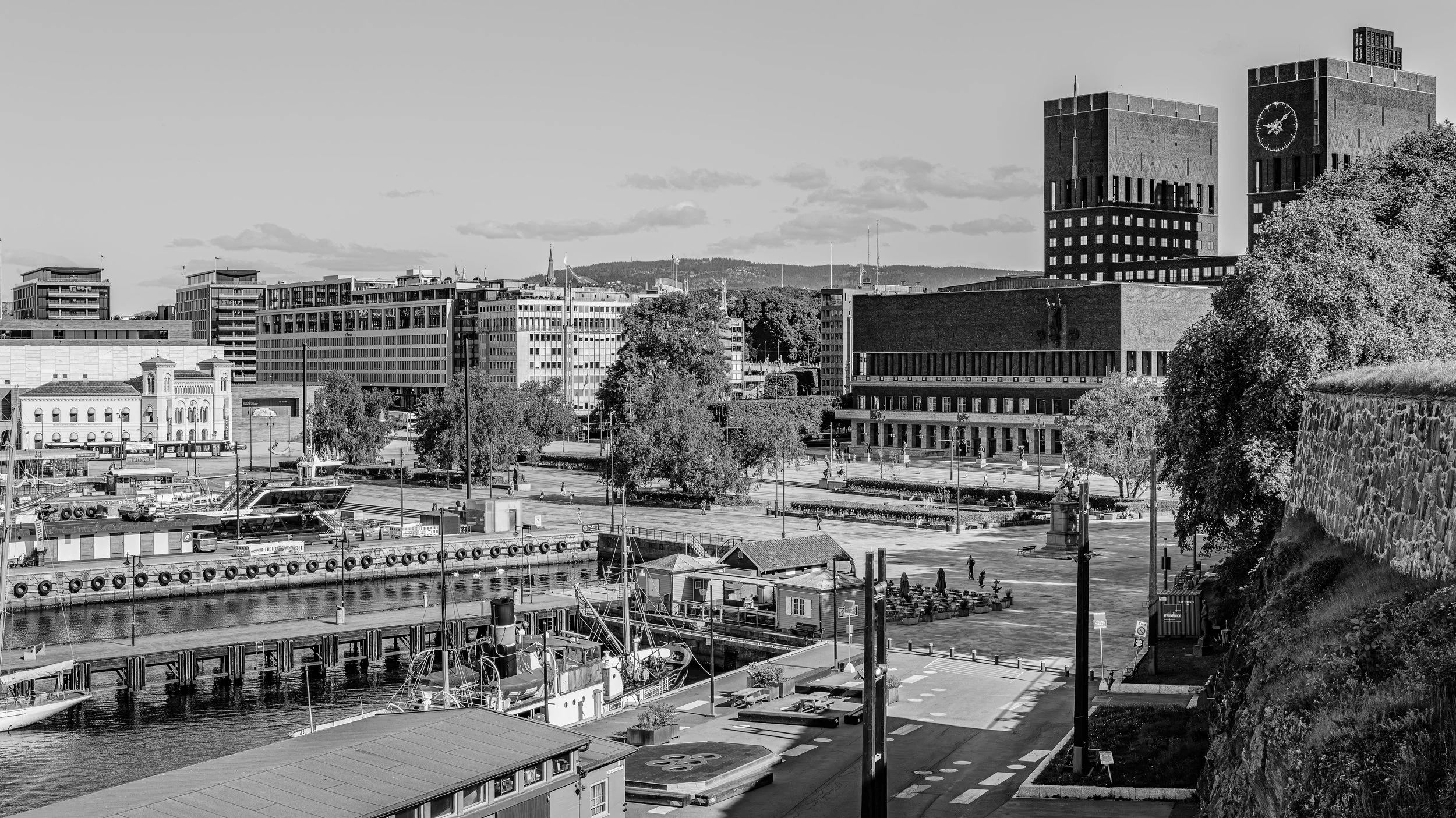 Black and white photo of a city skyline with modern buildings, a clock tower, trees, a waterfront with boats, and a promenade with people walking.