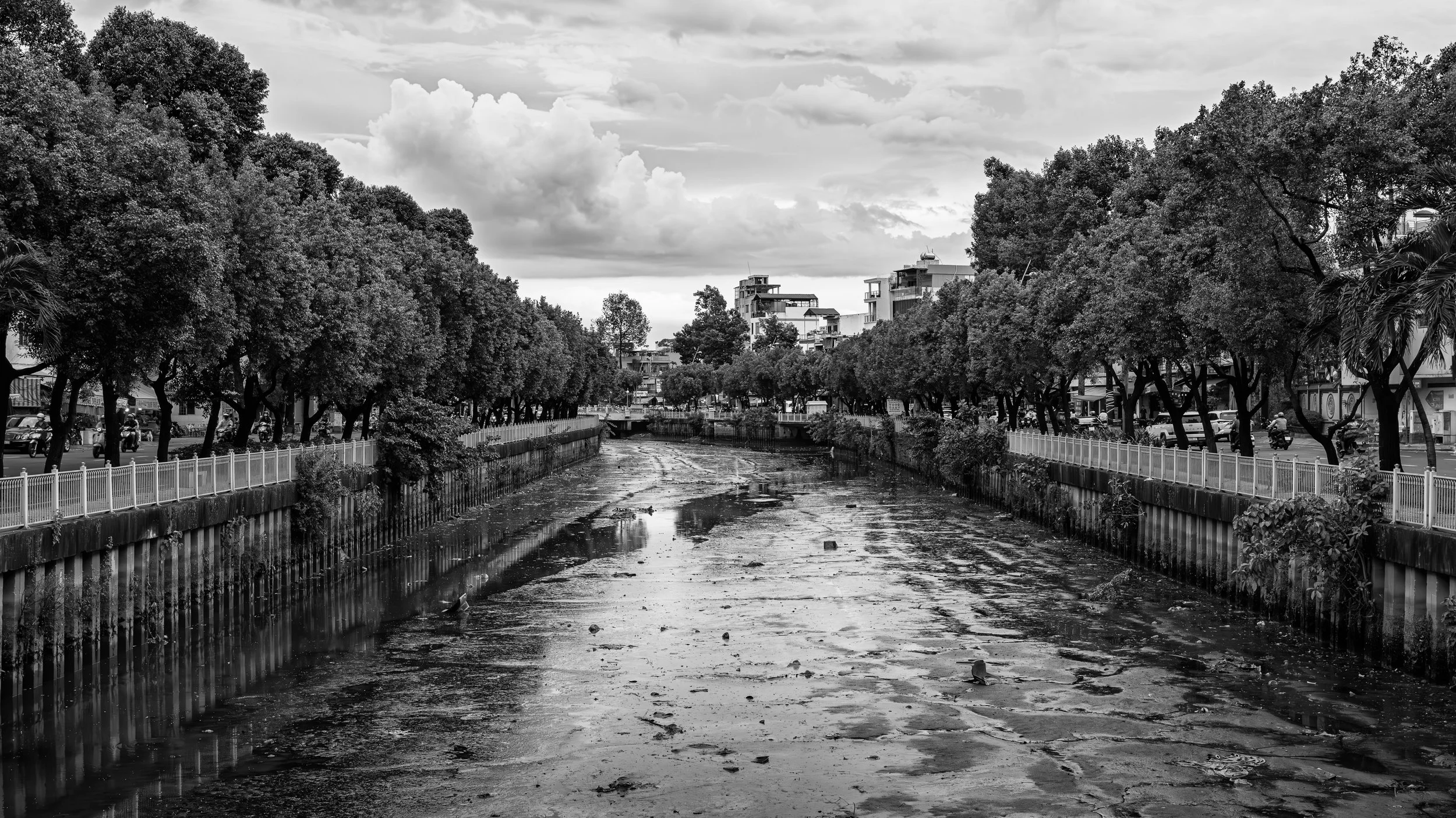 Black and white photo of a muddy, drained canal with debris, lined by trees on both sides, with residential buildings in the background and cloudy sky overhead.