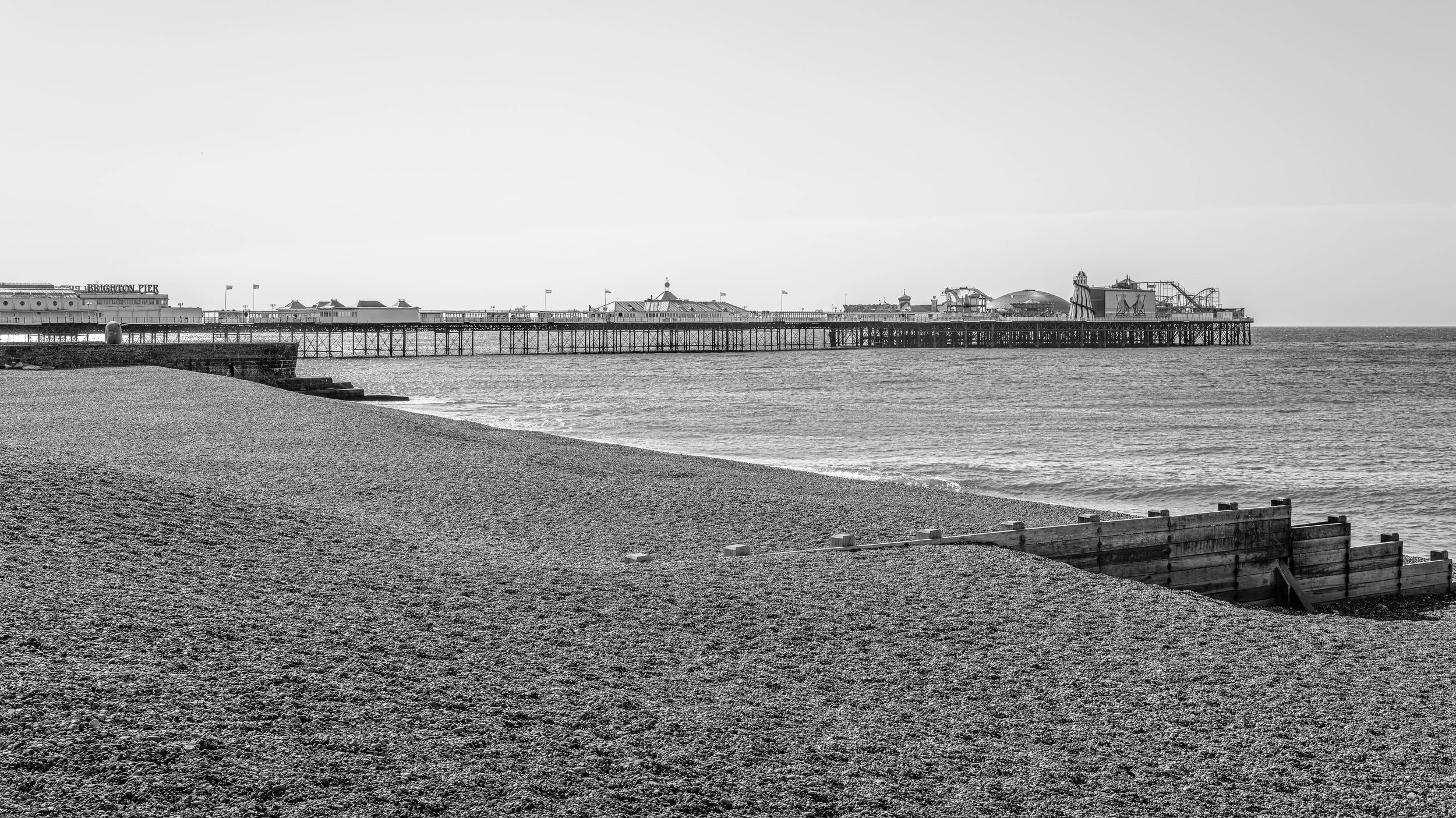 Black and white photo of a pier extending into the ocean with amusement park rides visible at the end, a beach in the foreground, and the sky above.