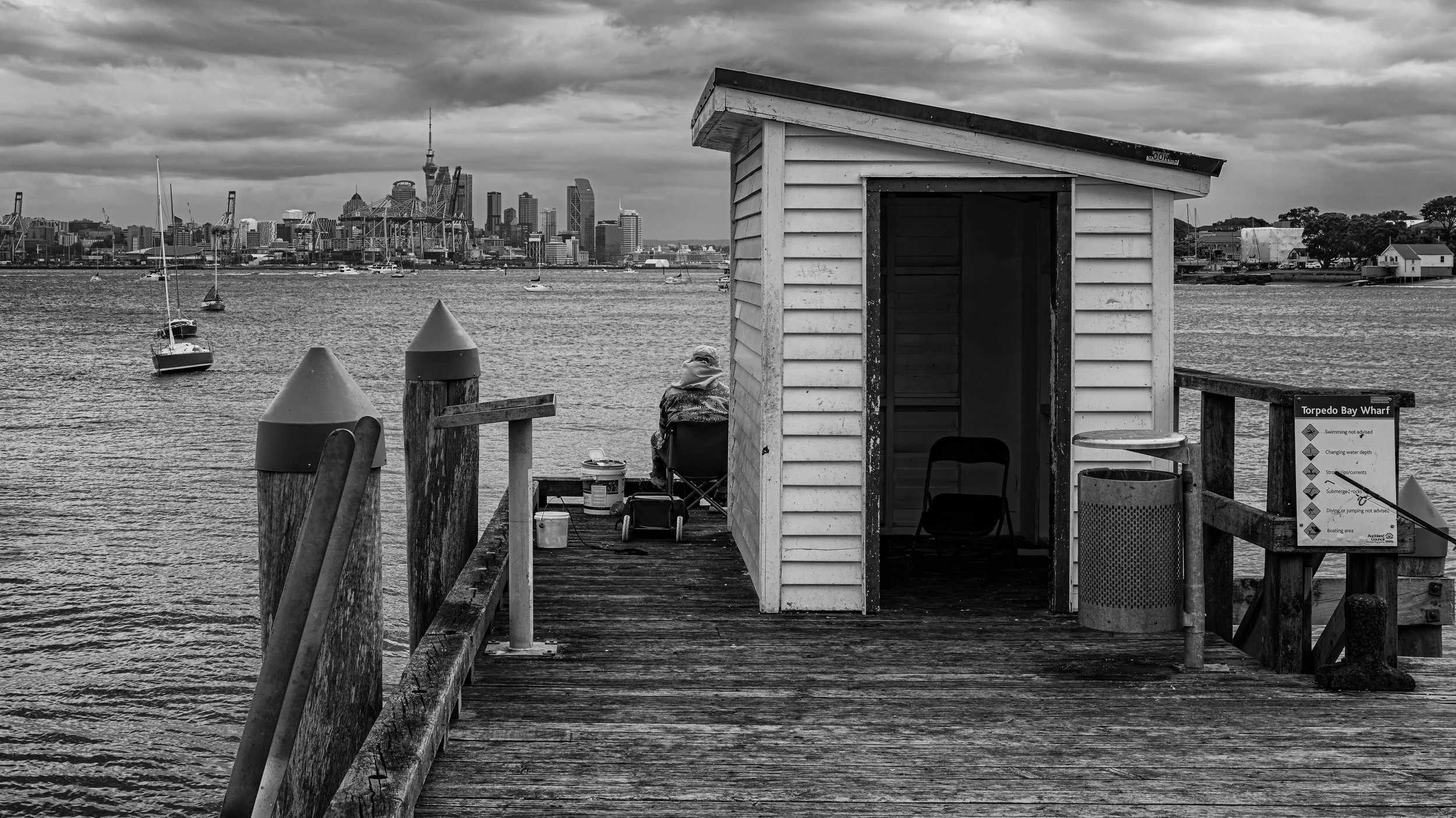 A pier at Torpedo Bay Wharf with a small wooden shelter, a person sitting on a chair, and a city skyline in the background on a cloudy day.