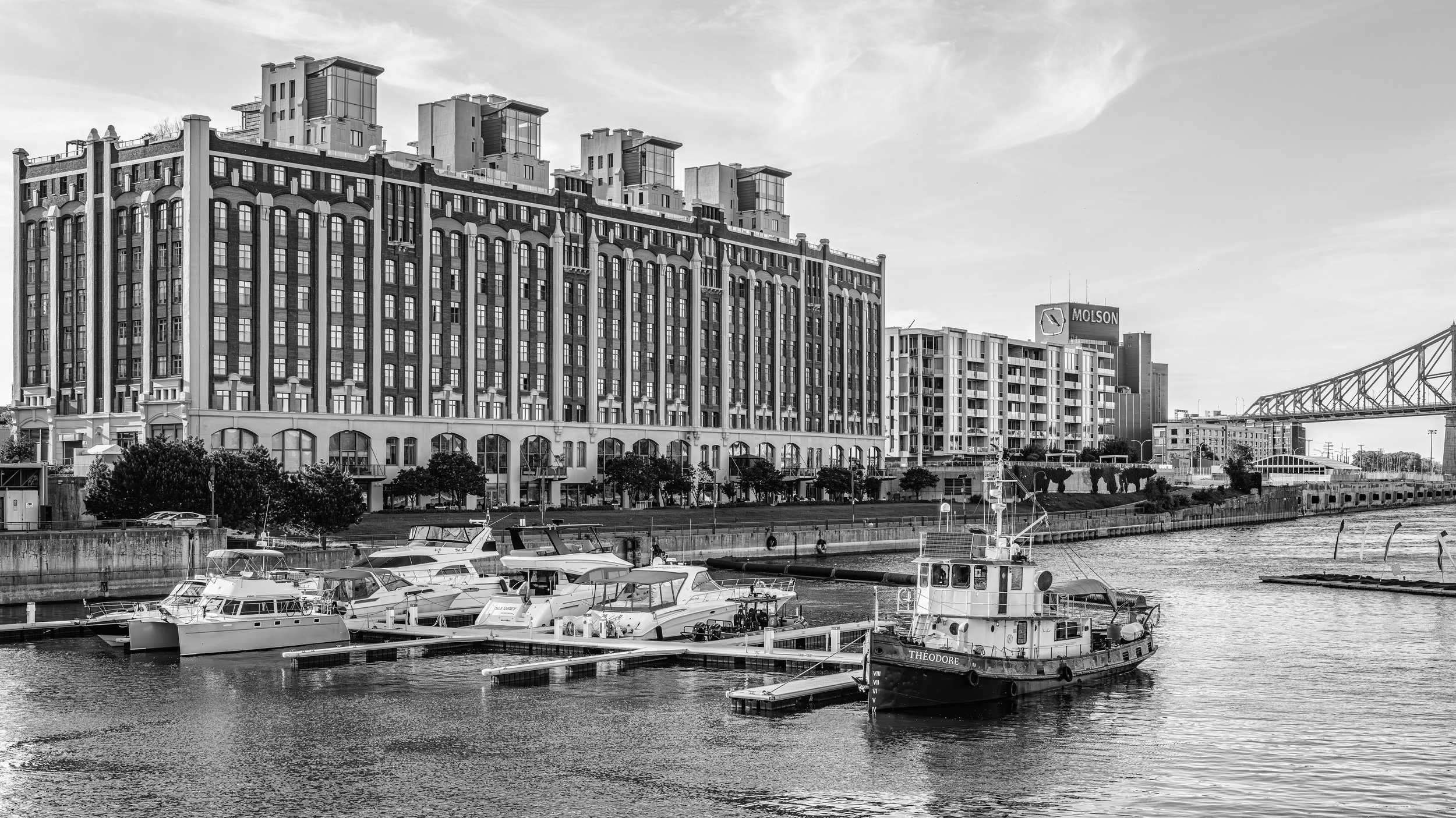A harbor with boats docked along a concrete pier, with modern multi-story buildings and a bridge in the background under a cloudy sky.