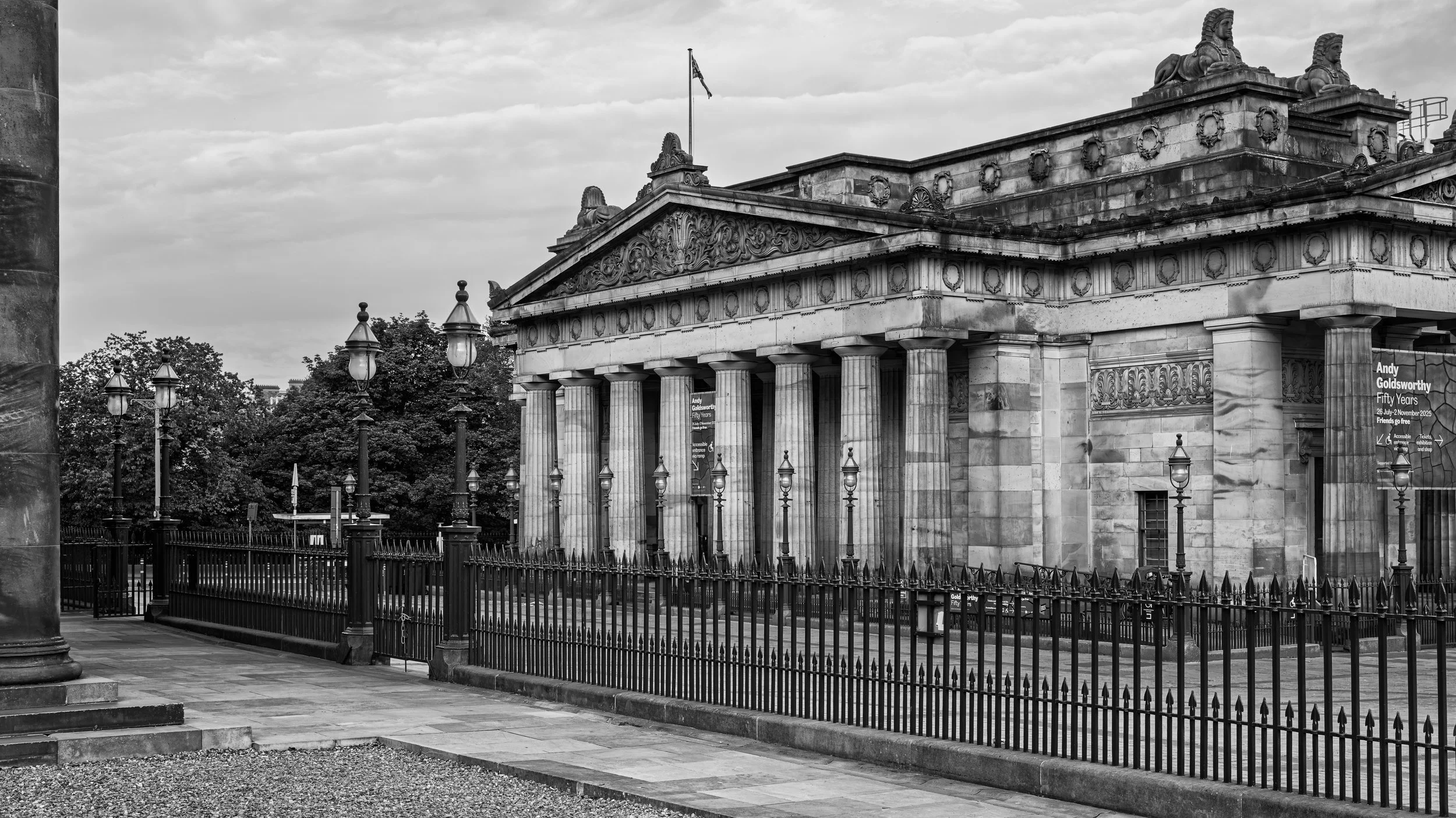 A black-and-white photograph of a classical building with Greek-style columns and detailed decorative elements on the pediment. There are street lamps and a gated fence in the foreground, trees in the background, and a cloudy sky overhead.