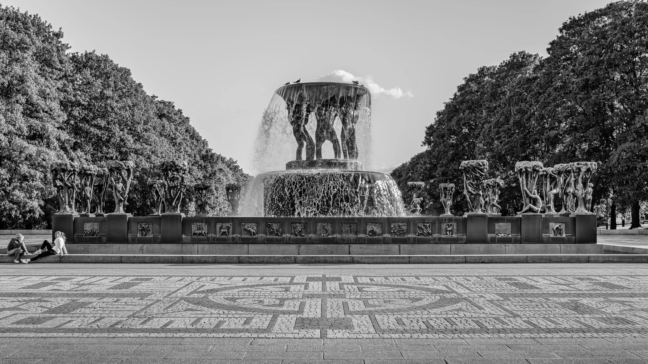 A large fountain with sculptures of animals and human figures surrounded by trees, with two people sitting near the base.