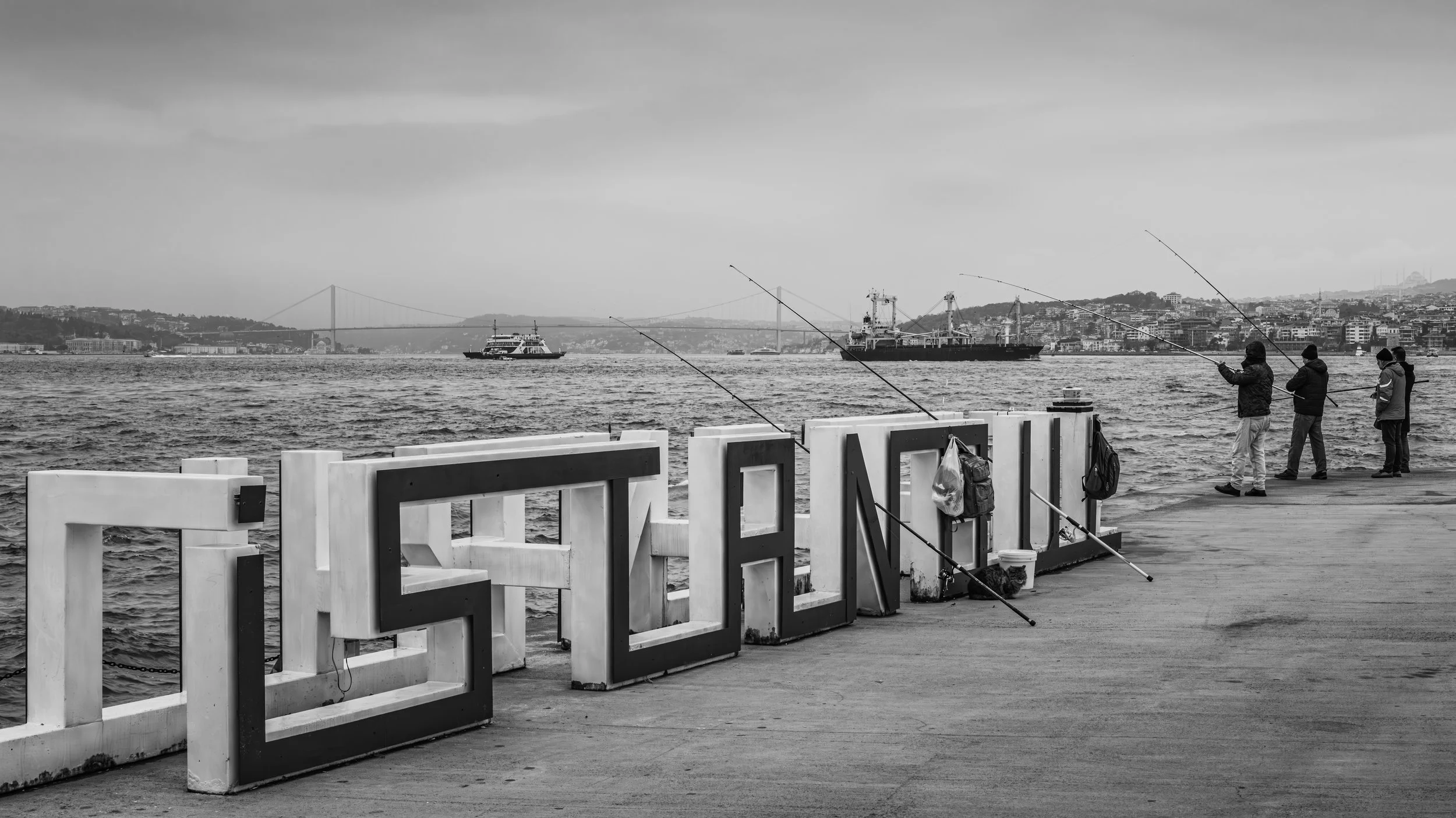 Four people fishing on a pier with boats in the water and a cityscape in the background, black and white photo.