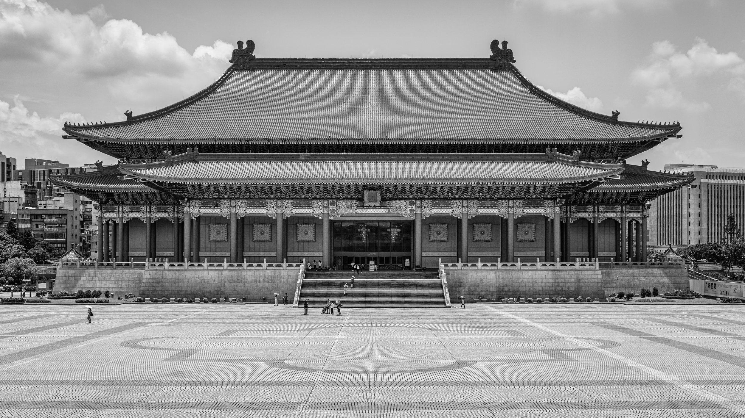 A traditional Asian-style building with ornamental roofs and intricate details, set against a partly cloudy sky, with a wide paved plaza and few people walking in front.