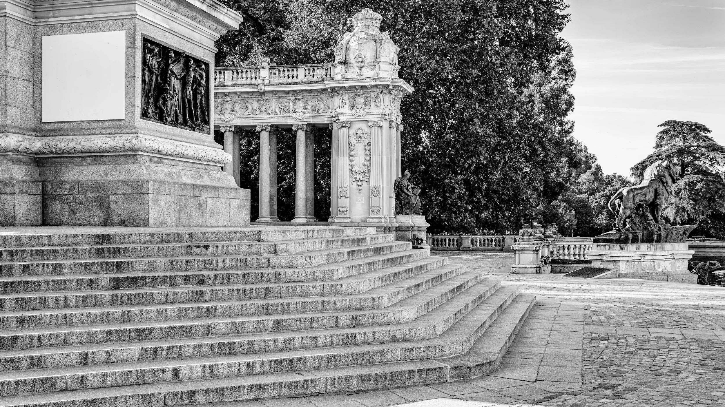 A black and white photo of a historical monument with stairs leading up to it, accompanied by statues of horses and figures, surrounded by trees in the background.