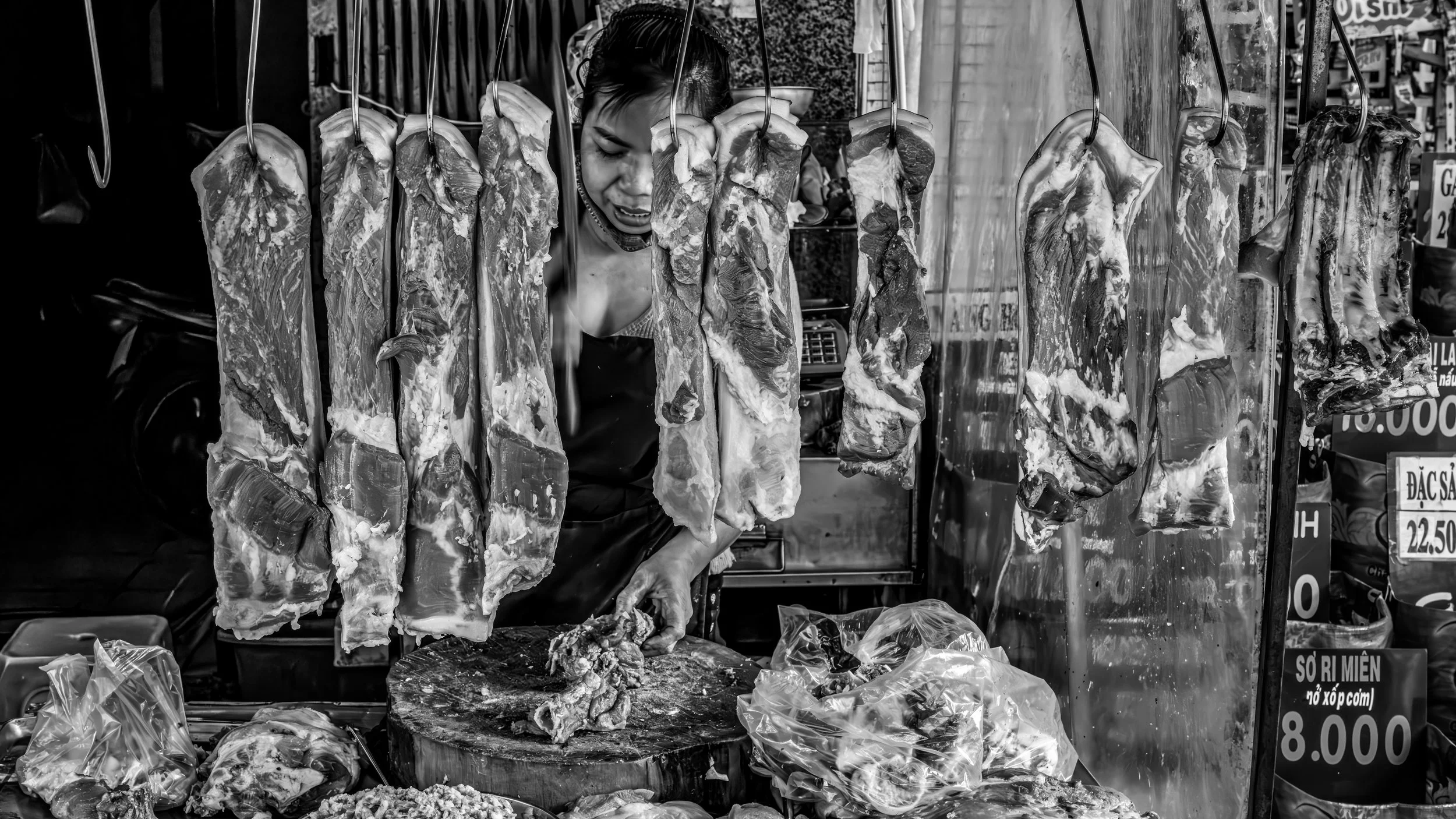 Black and white photo of a woman at a market stall, butchering and selling large cuts of meat hanging from hooks.