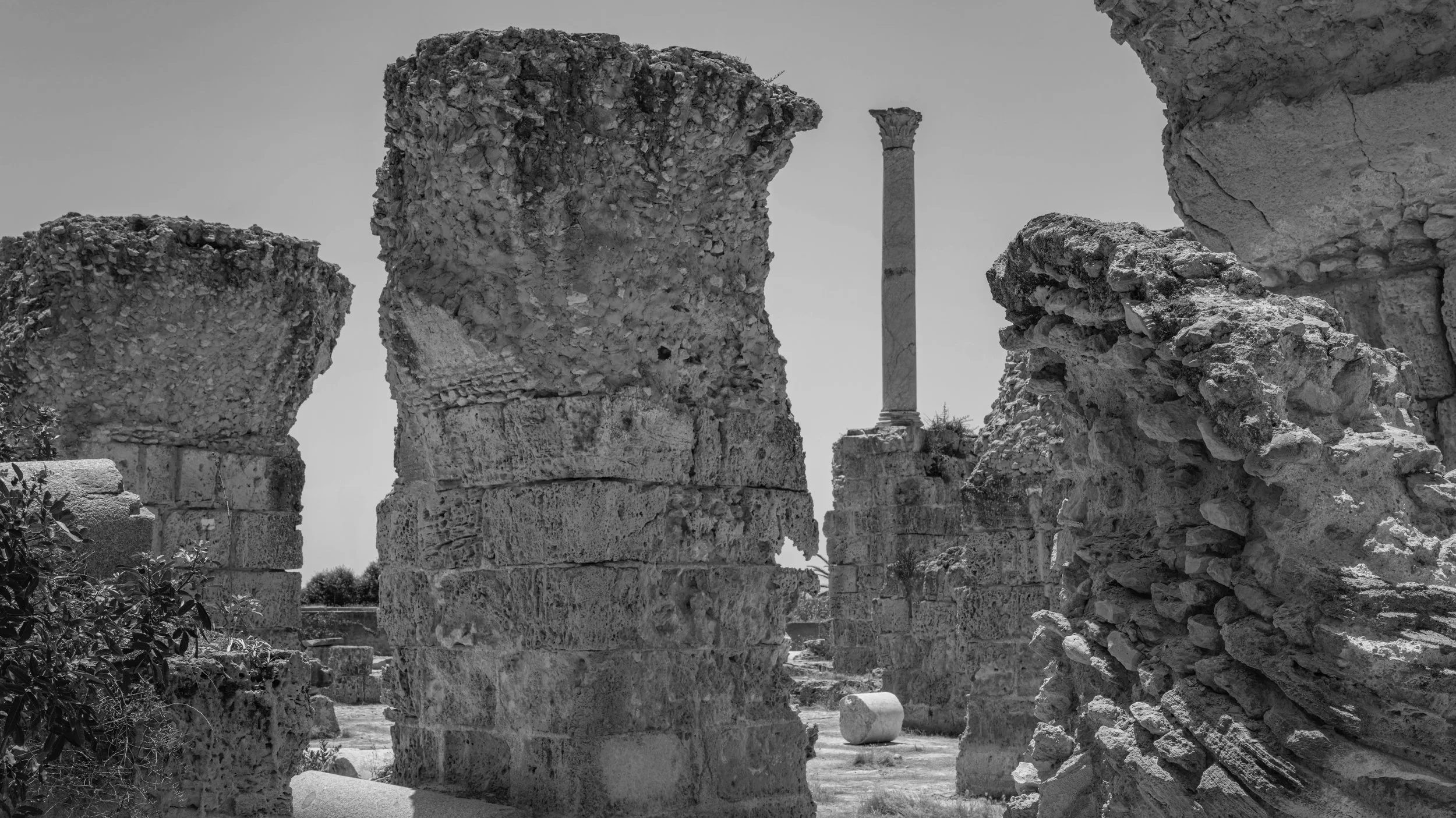 Ancient ruins with stone columns and broken structures, with a tall column in the background, under a clear sky.