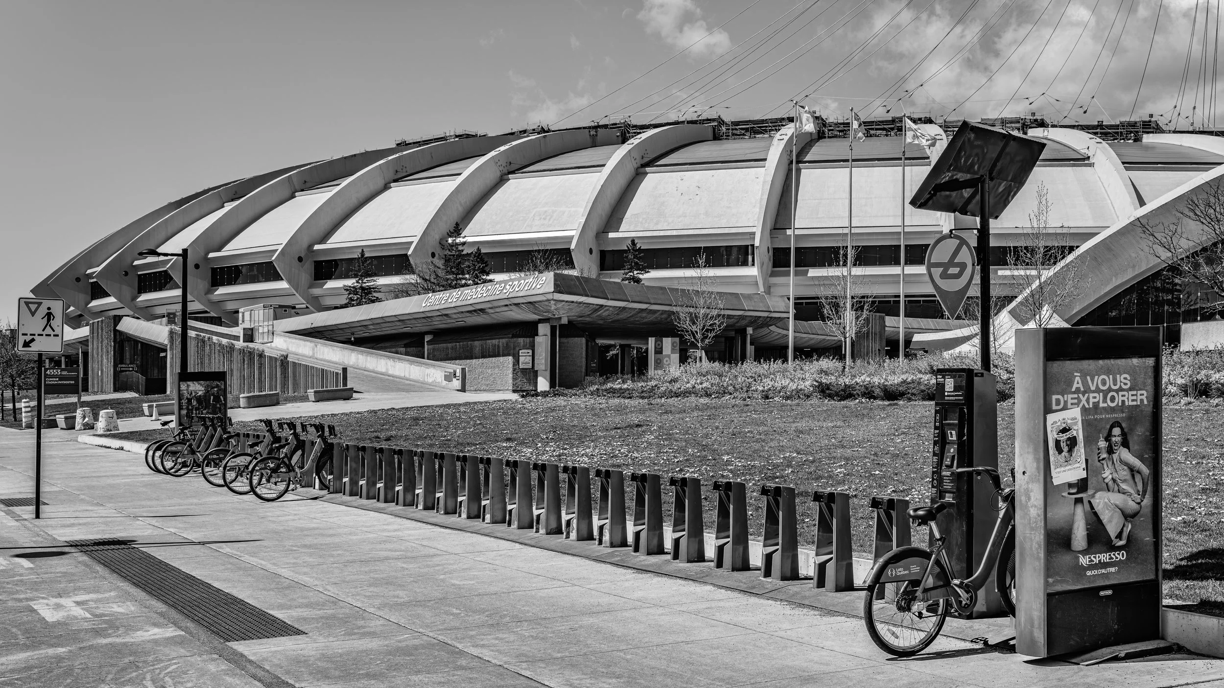 Black and white photo of the Stade de France stadium. There are bicycles parked on the sidewalk, a bus stop with an advertisement, and a sign indicating pedestrian and vehicle pathways.