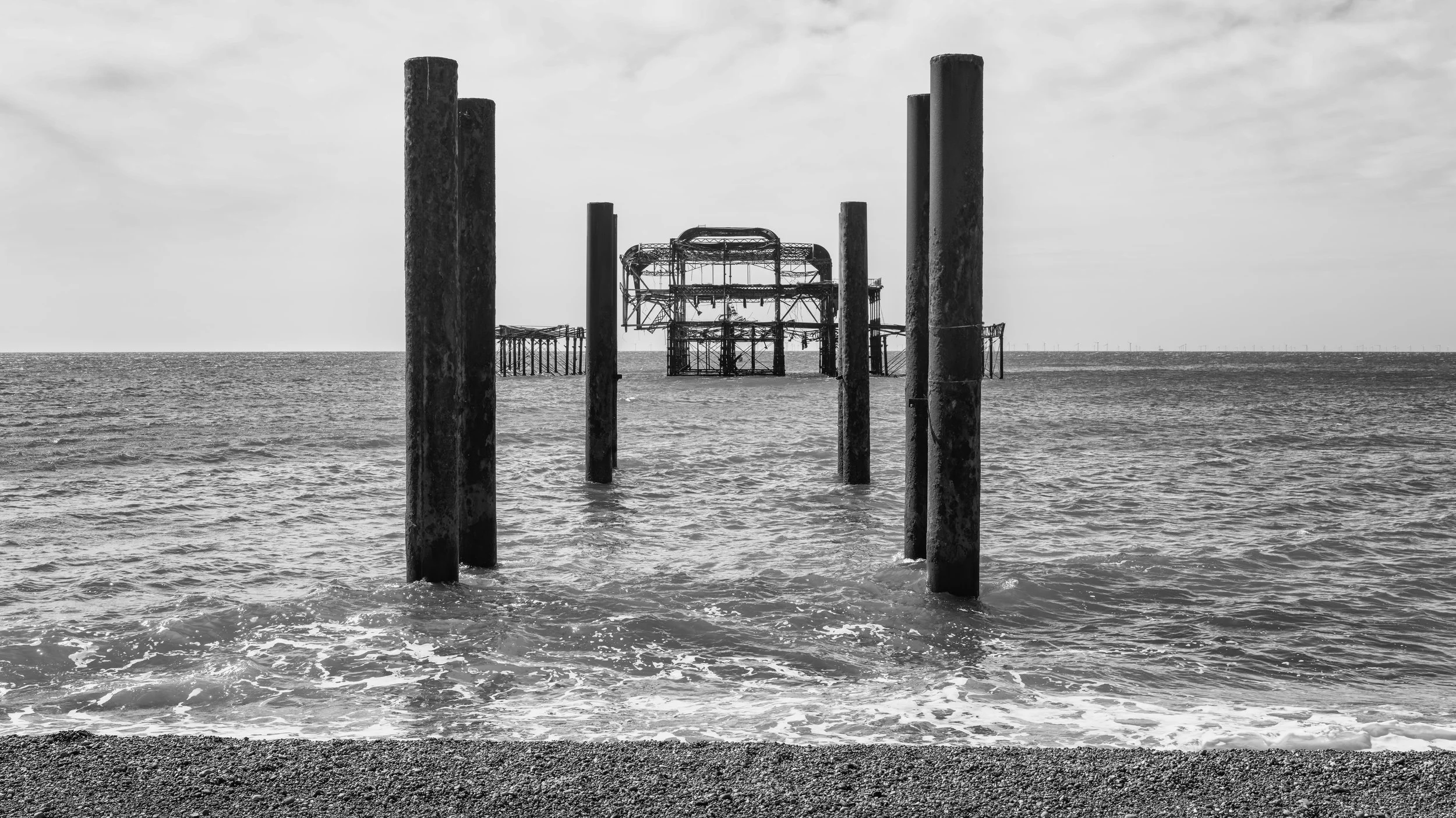 Black and white photo of remnants of an old pier with several weathered vertical posts in the water, and a dilapidated metal structure in the background, along the shoreline.
