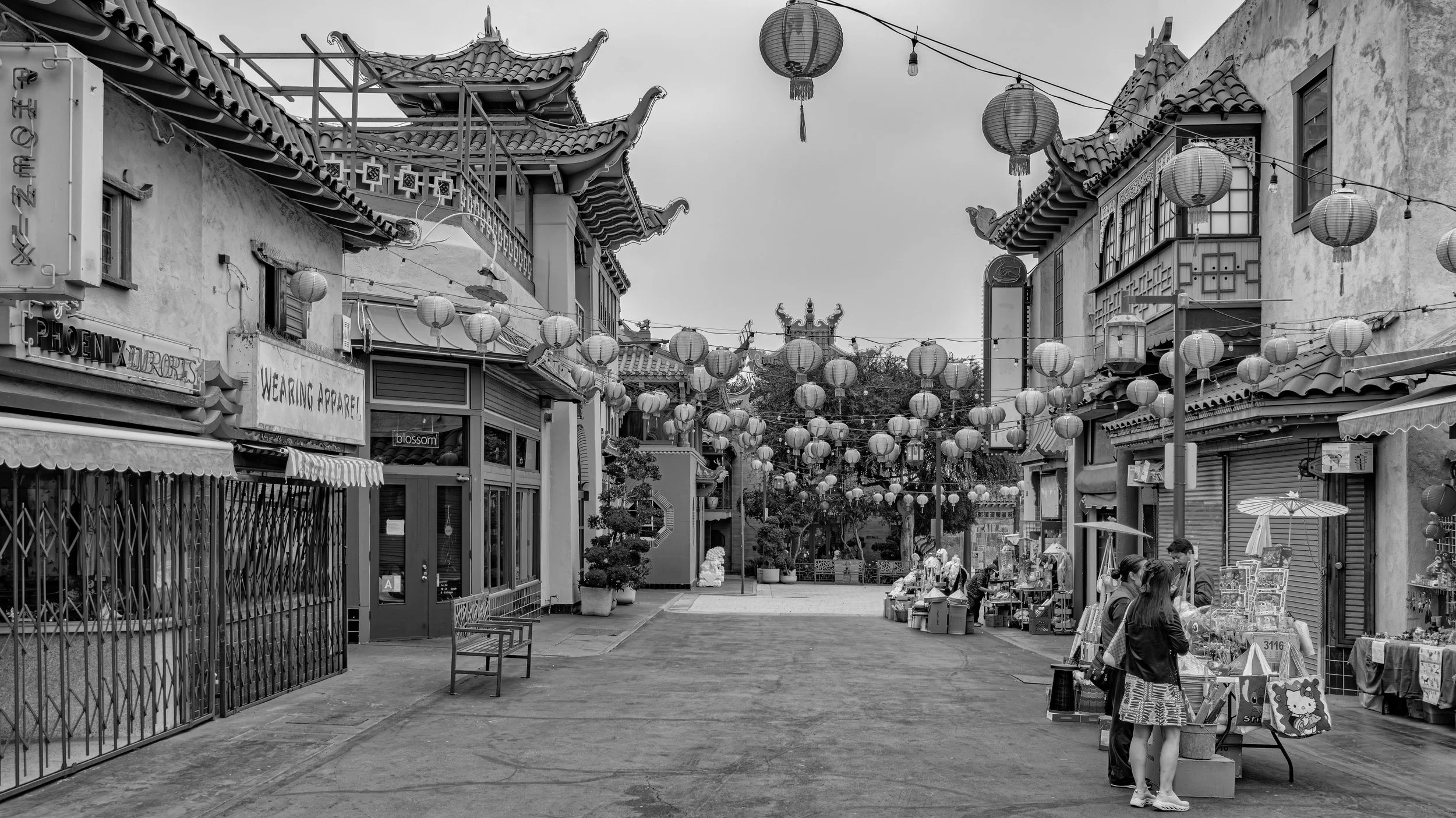 A street scene with Asian architectural elements, decorated with hanging lanterns. Shops and stalls line the street, with people browsing and walking. The sky is clear, and the scene appears lively and colorful.
