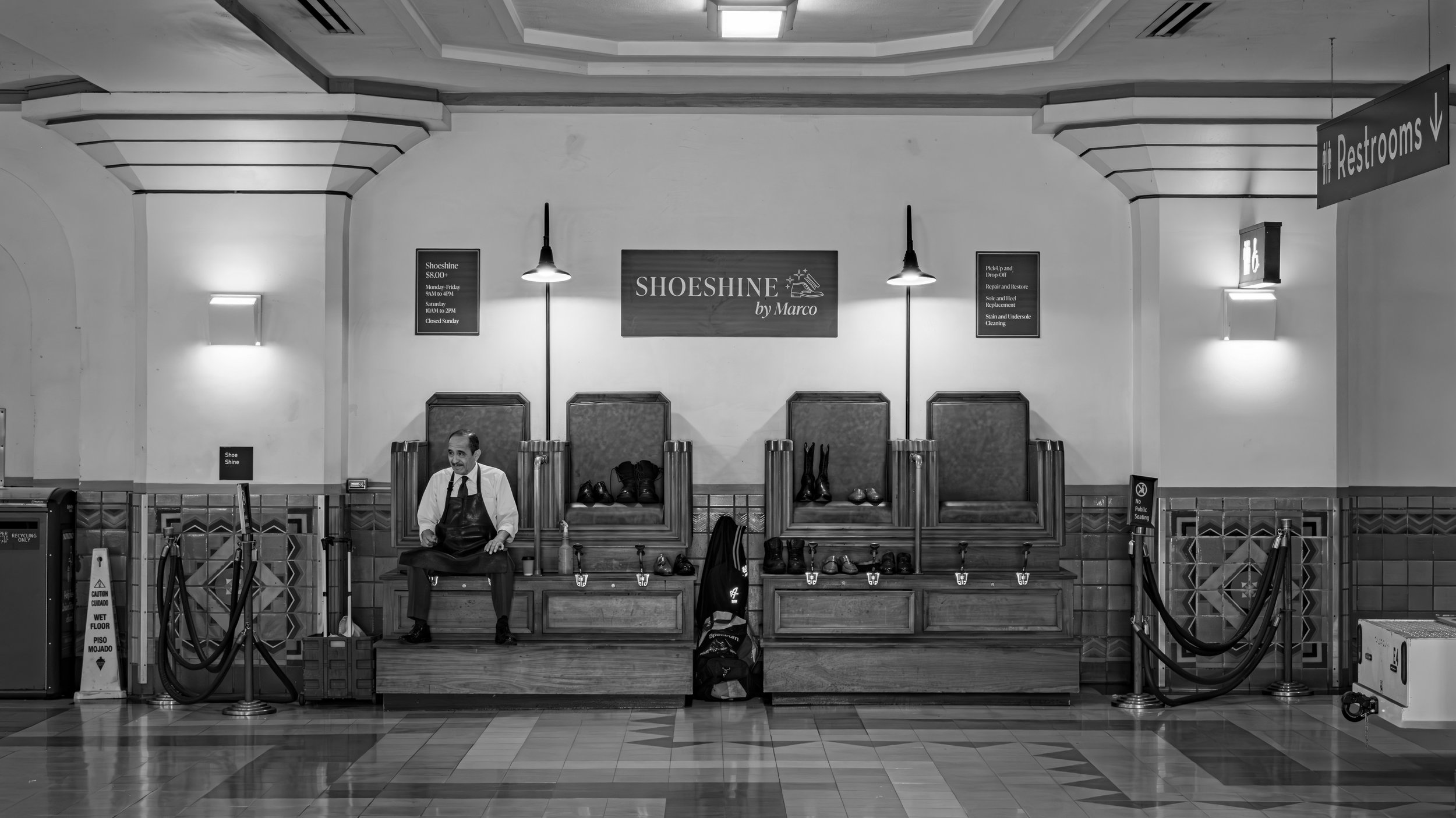 A man sits on a bench in front of shoe racks in a waiting area, with signs indicating shoe shine services and restrooms in the background.