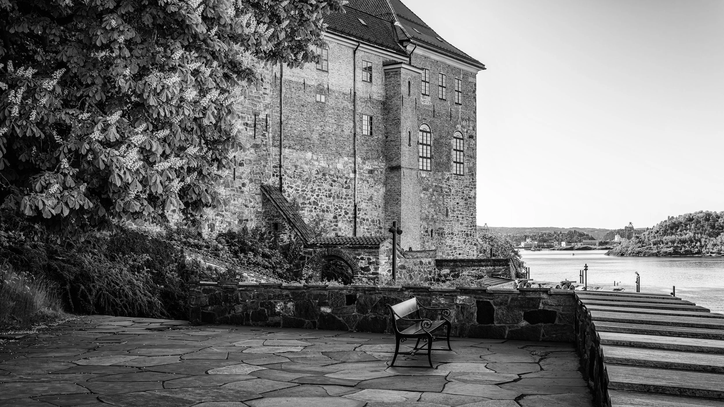 Black and white photo of a historic brick and stone building by a river with a tree on the left and a bench in the foreground.