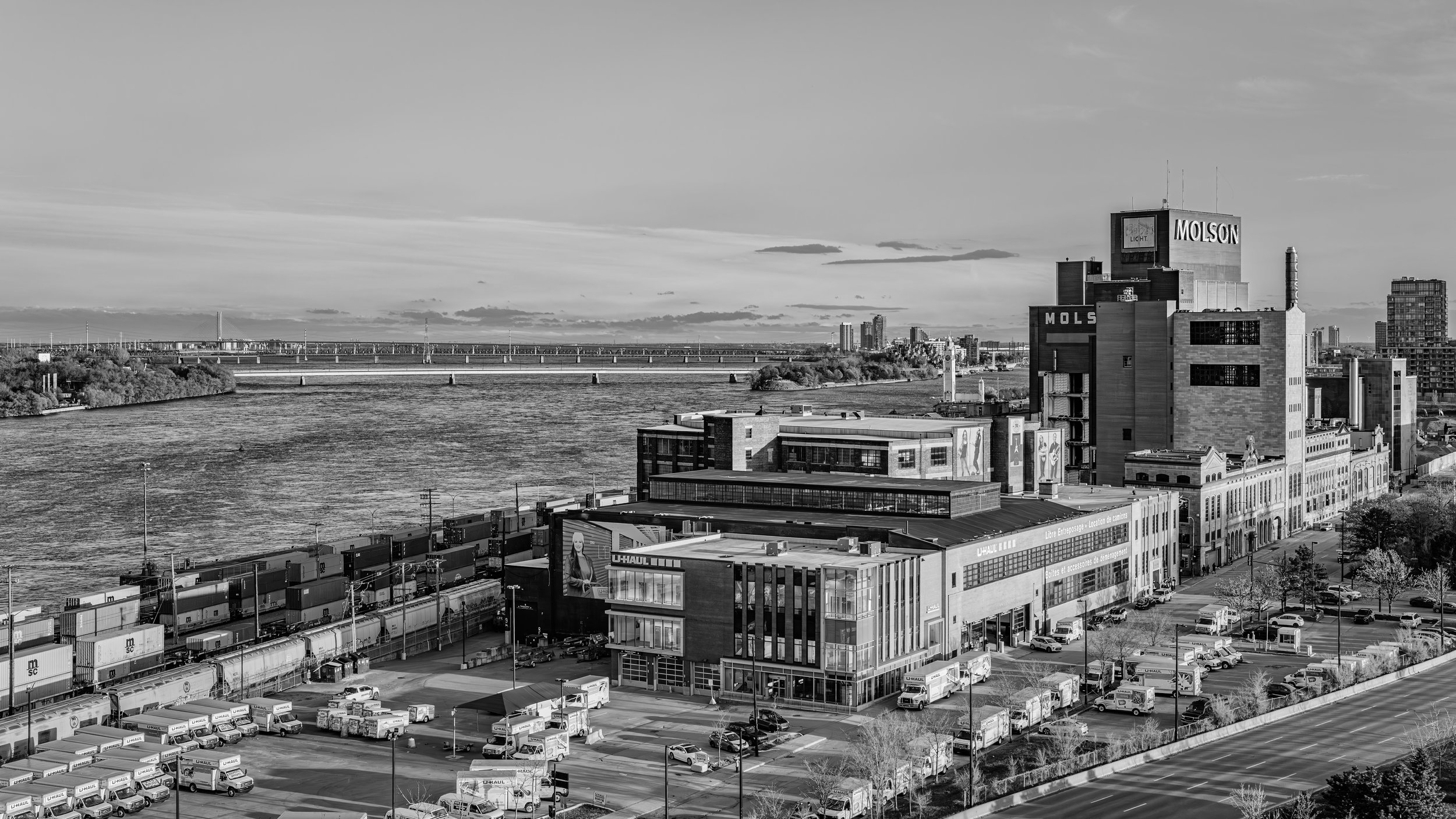 Black and white cityscape with a river in the background, industrial buildings, shipping containers, and a bridge crossing the river.