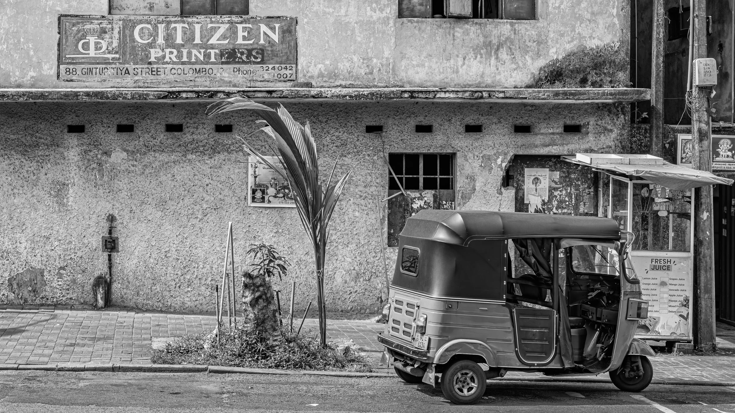 A three-wheeler auto rickshaw parked on the street near a small juice stall, with an old building in the background that has a sign for Citizen Printers on Ginturtiya Street in Colombo, Sri Lanka.