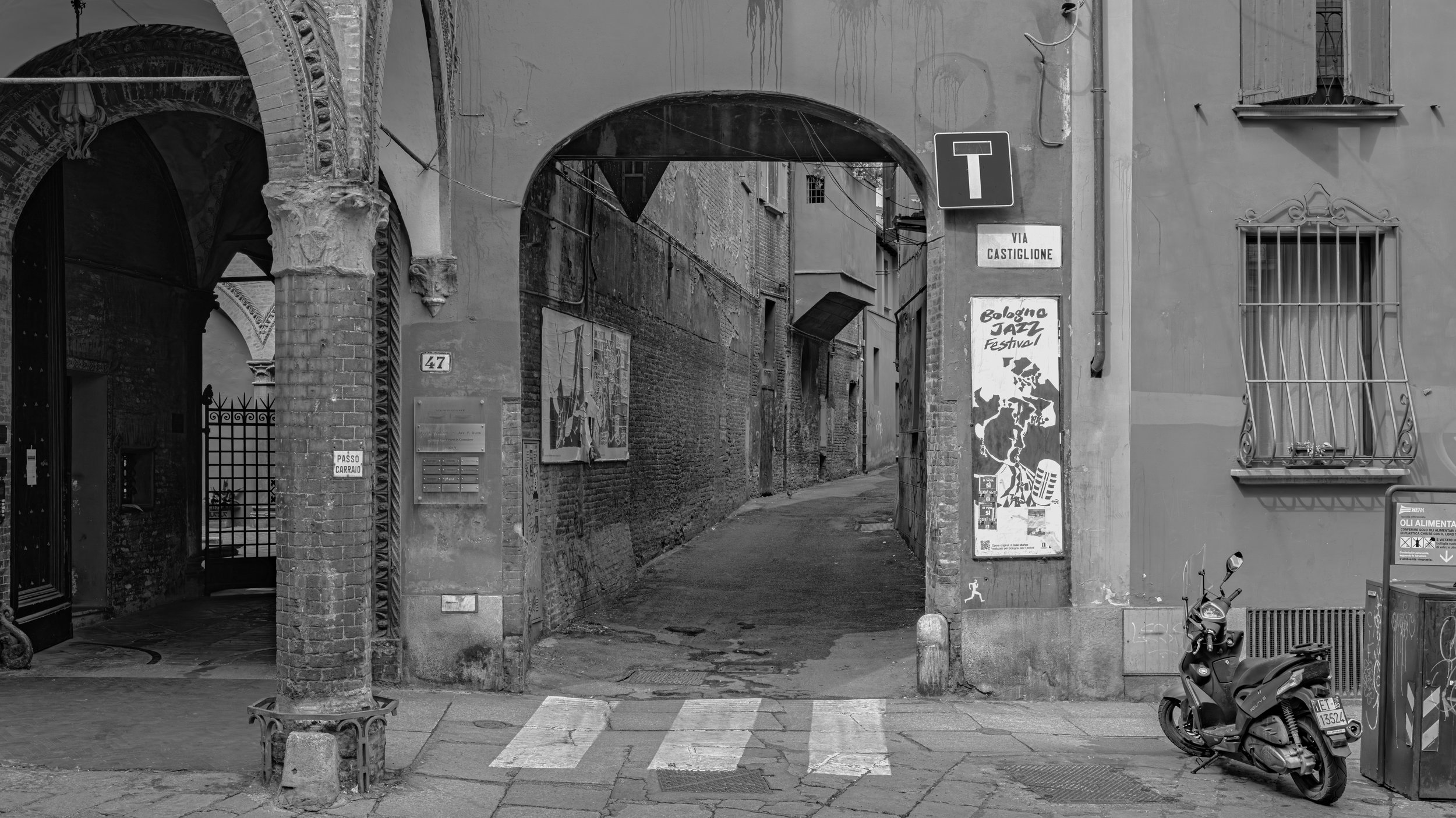 A narrow alleyway framed by an arch gate with graffiti, signs, and a small motorcycle parked outside in an old European city.