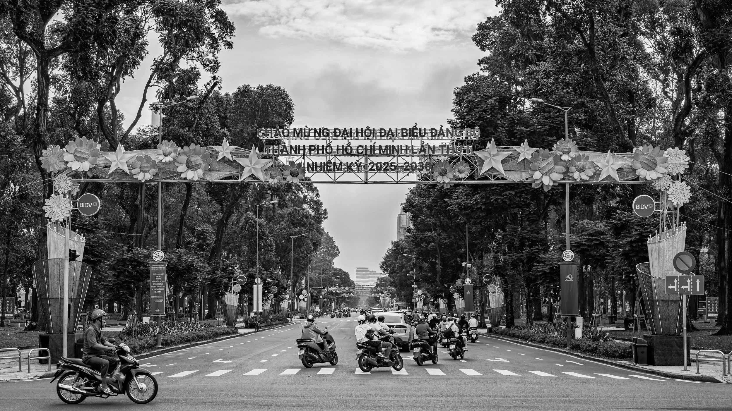 Street scene with motorbikes and cars, large decorative archway with flowers and stars, and trees lining the street in Hanoi, Vietnam.