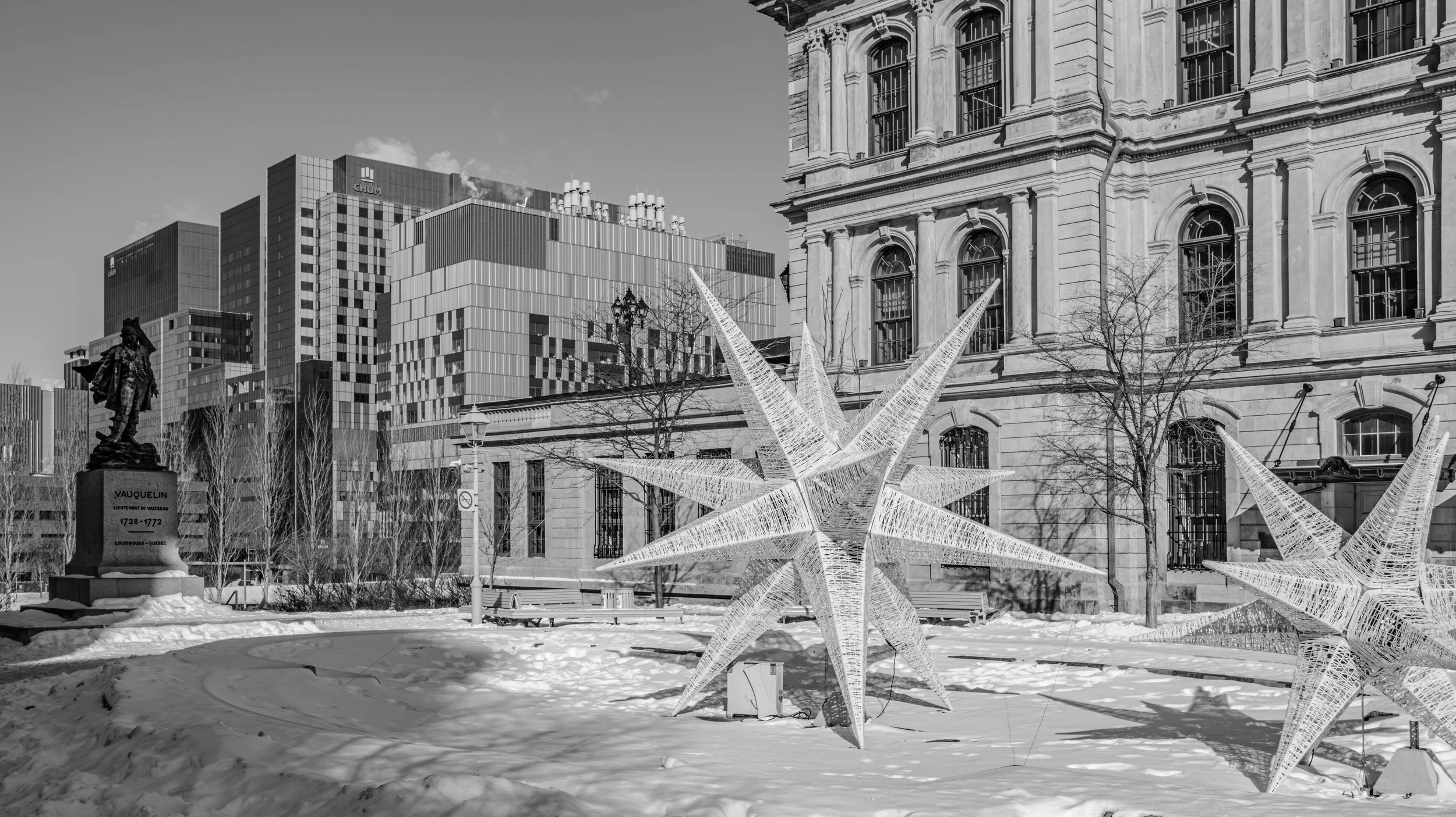 Black and white photo of an urban winter scene with buildings, a statue, leafless trees, and illuminated star decorations on the snow.