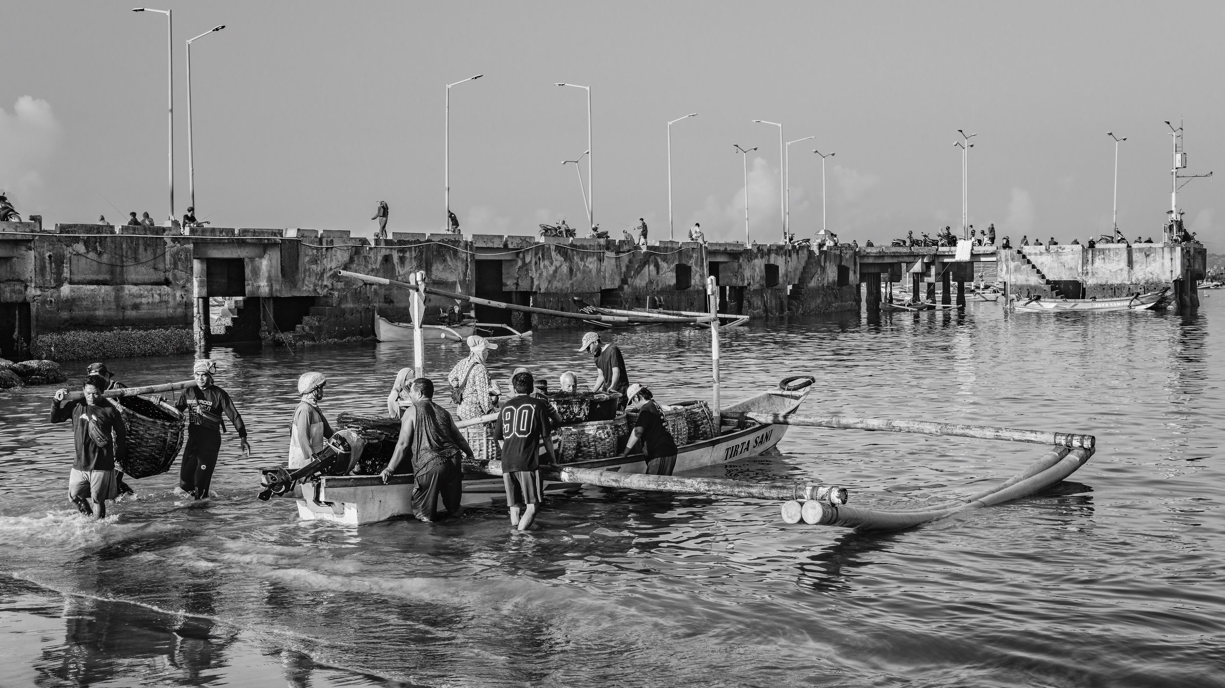 Black and white photo of people working on boats by a pier, some with fishing equipment, in a body of water with a concrete pier and power lines in the background.
