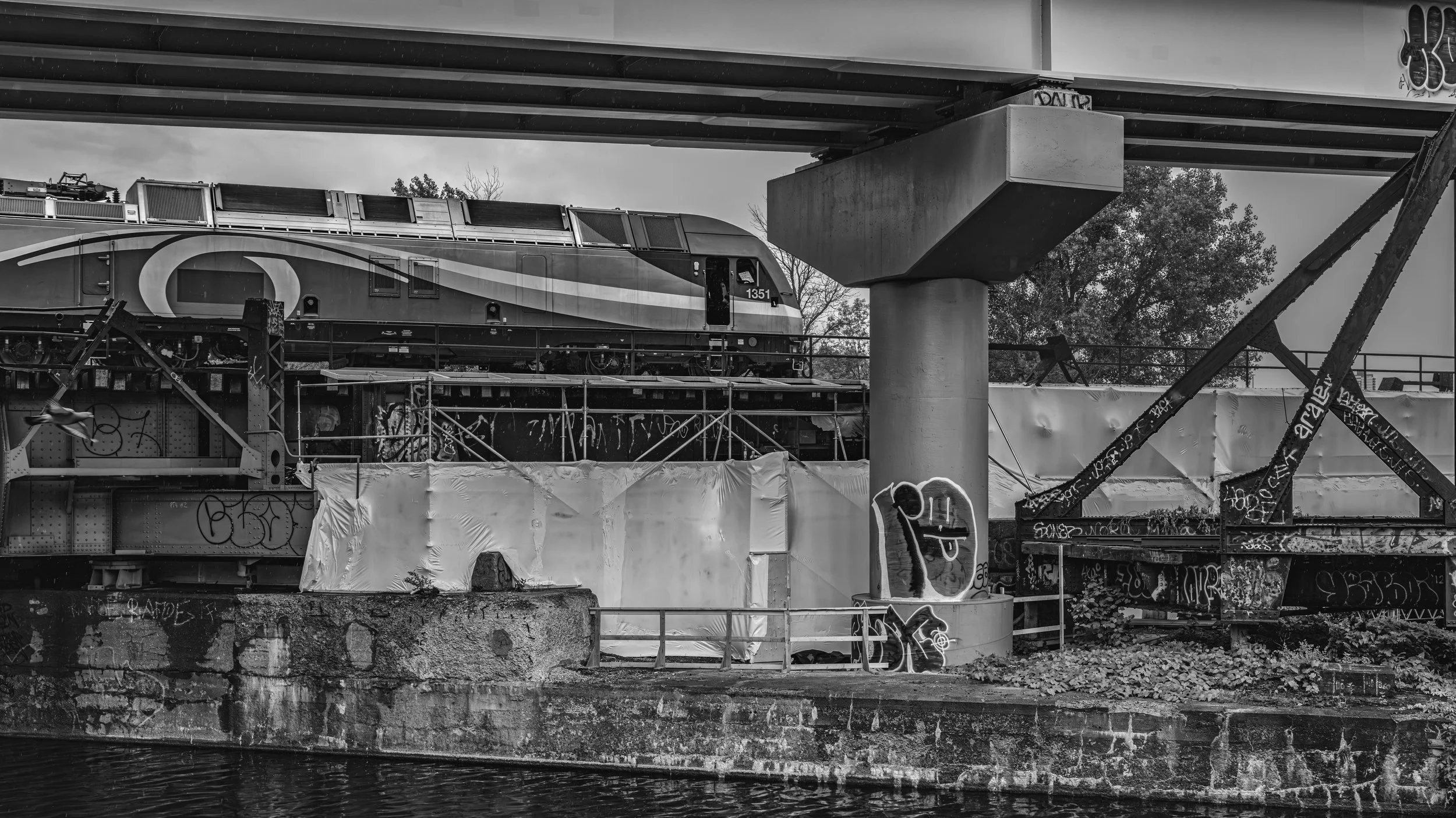 Black and white photo of a train on a bridge under construction, with graffiti on the support beams, and a water body at the bottom.