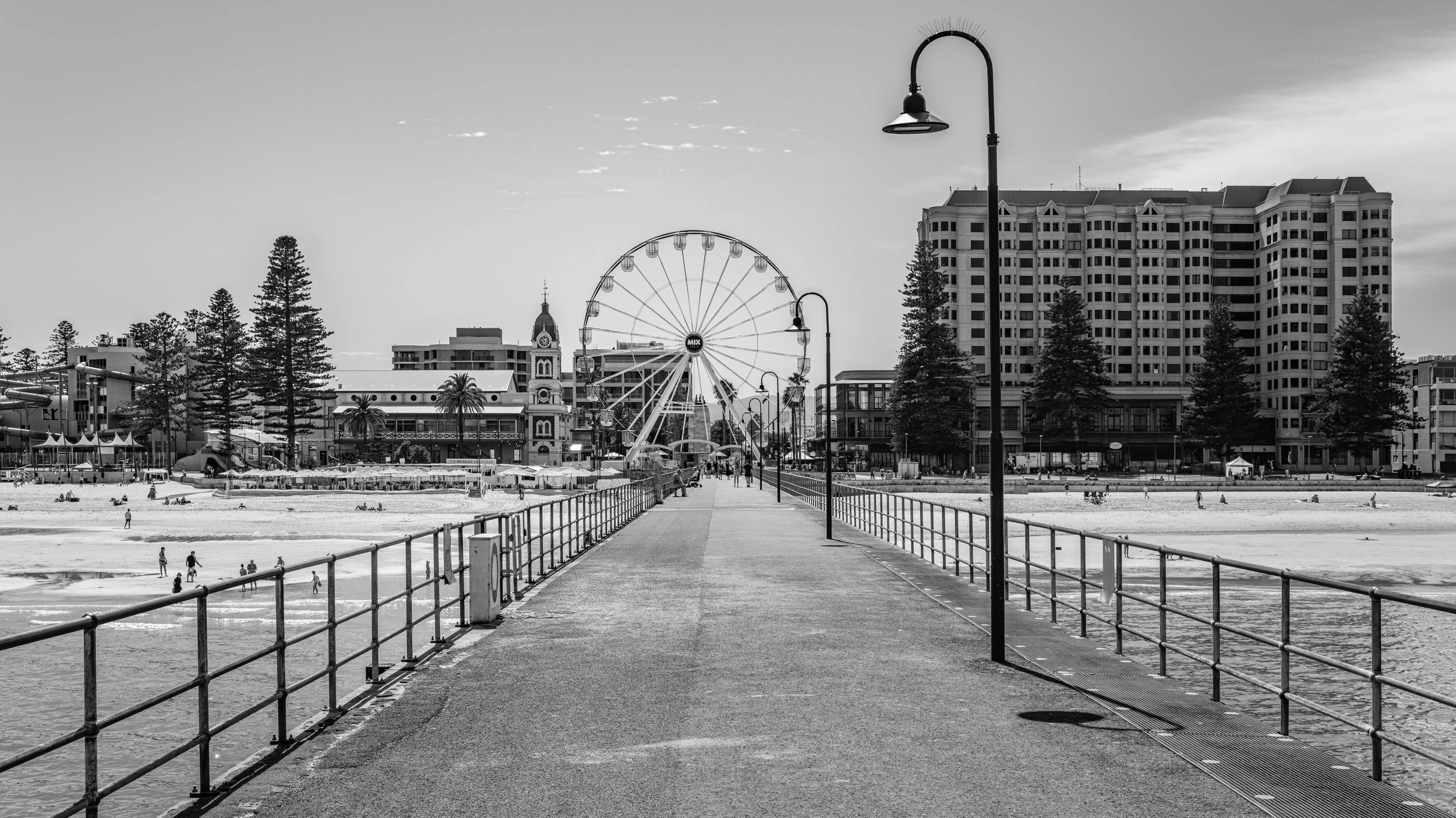 A black-and-white photo of a pier extending into a beach with a city skyline in the background, including a ferris wheel and tall buildings.